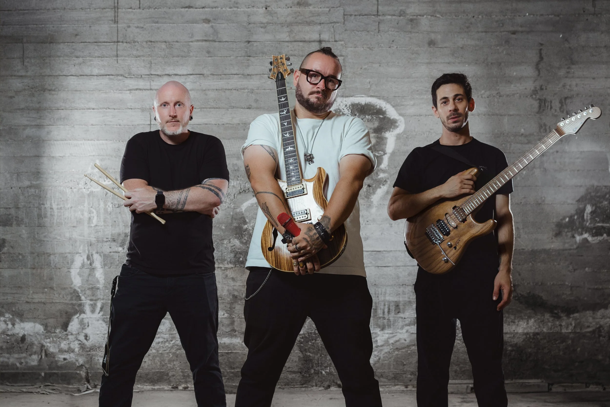 Three men posing with musical instruments against a concrete wall. The man in the center holds an electric guitar and has tattoos and a beard. The man on the left holds drumsticks and has a shaved head. The man on the right holds an electric guitar a