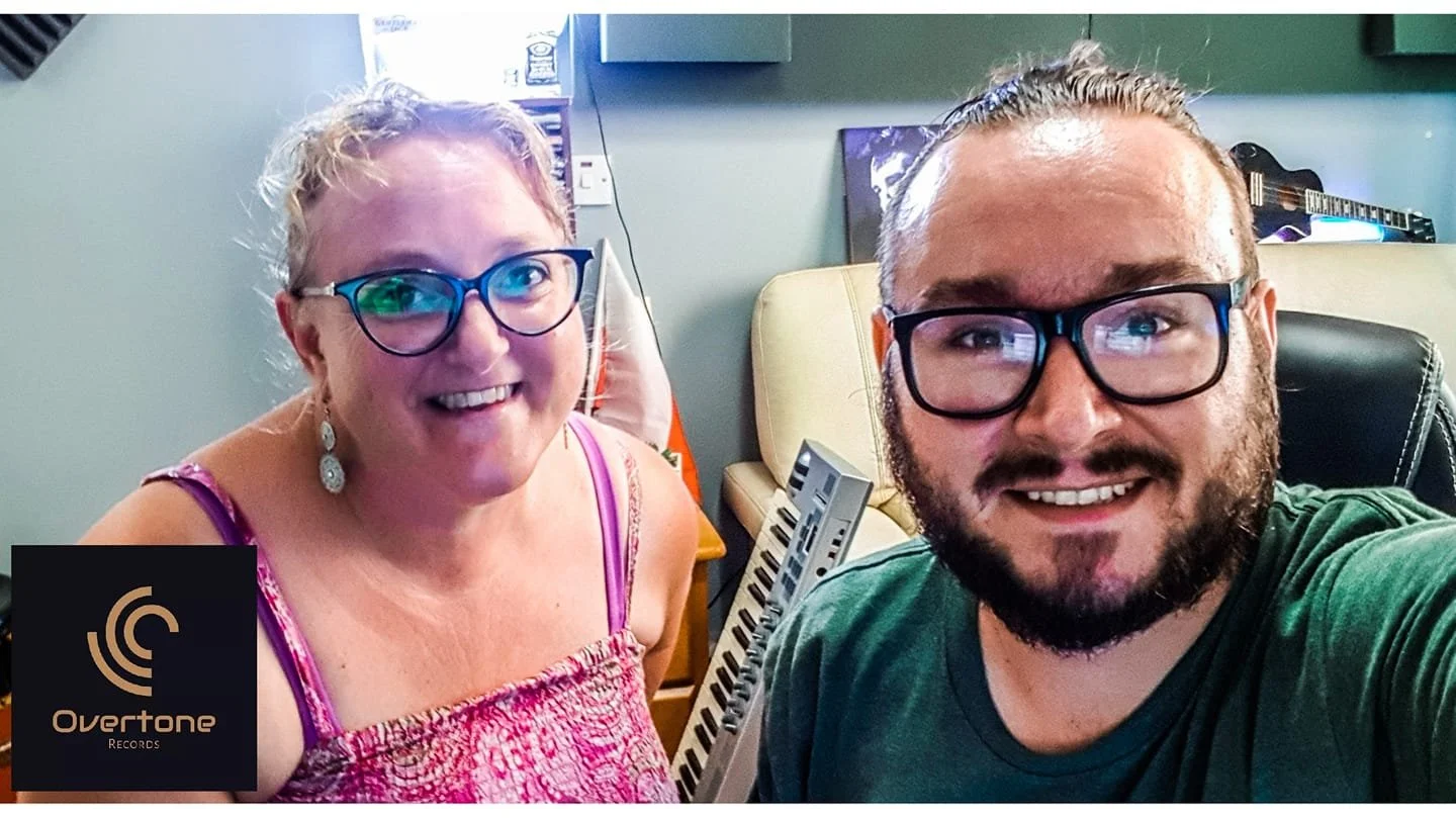 A woman and a man taking a selfie in a music studio. The woman has glasses, earrings, and a pink top, and the man has glasses and a beard. There is musical equipment and a logo for Overtone Records in the background.