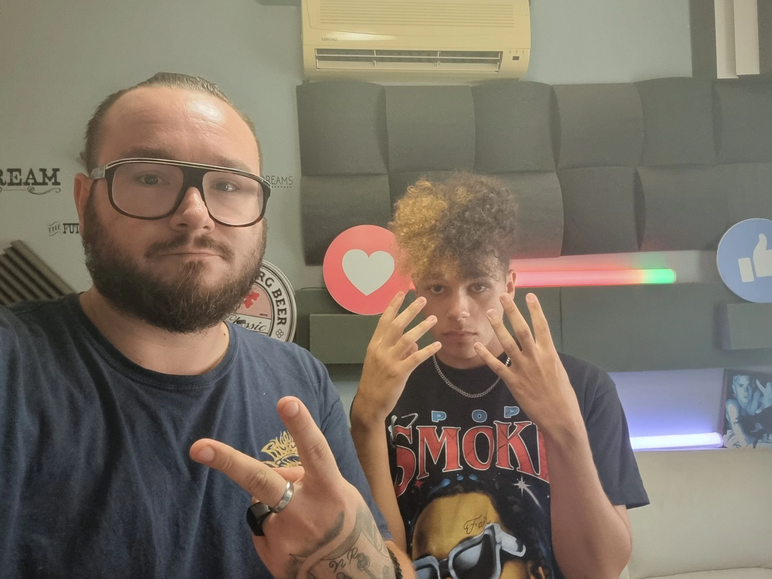 Two men taking a selfie indoors. The man on the left has glasses, a beard, and tattoos. The man on the right has curly hair and is holding up his hands with fingers spread. There are wall decorations and an air conditioning unit above them.
