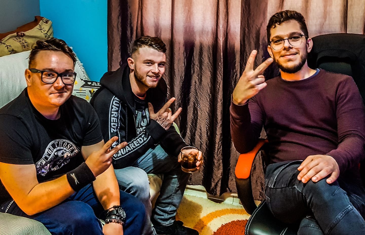 Three young men sitting together in a living room, smiling and making hand gestures. One is seated on a white television bench, the second on a chair, and the third on an orange chair. The middle man has a drink in hand. They are casually dressed, wi