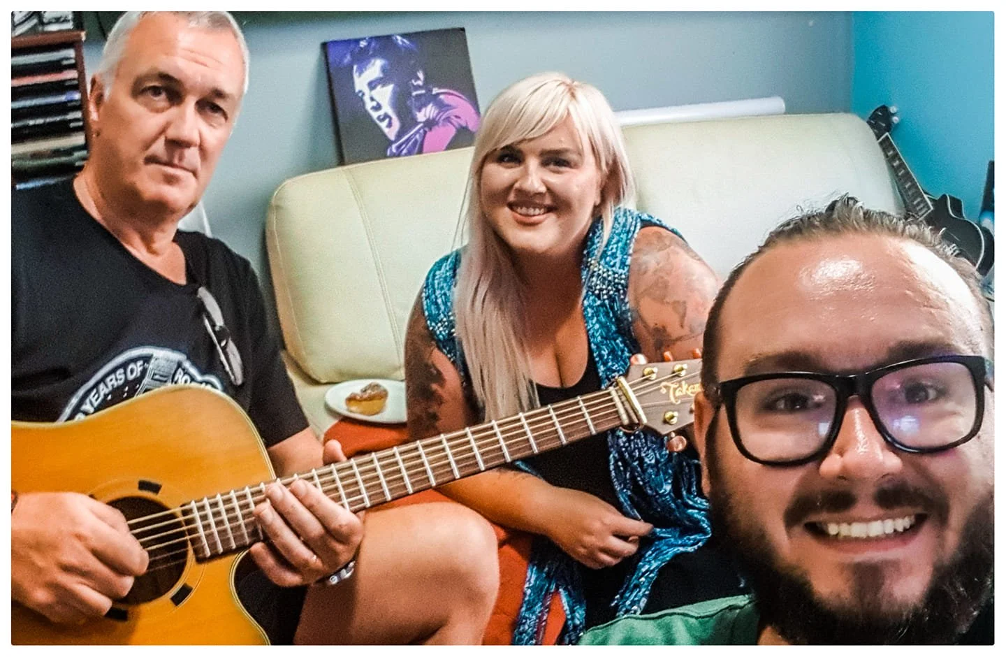 Three people in a room, one playing guitar, smiling and looking at the camera. The woman in the middle has blonde hair, tattoos, and is smiling. The man on the left has gray hair and is also smiling. The man on the right has glasses, a beard, and is 