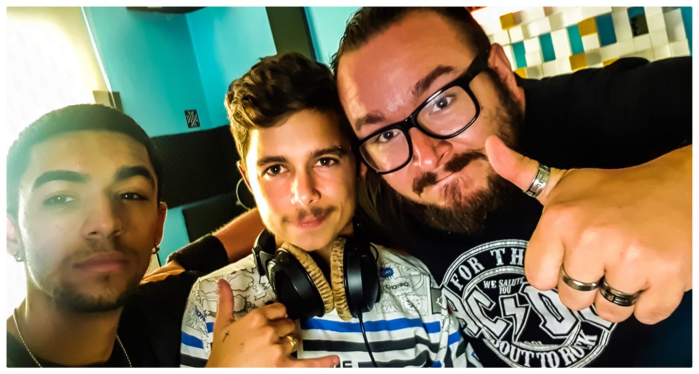 Three men taking a selfie together in a brightly lit indoor setting, with blue walls and colorful decor in the background. They are close together, smiling and making gestures with their hands.