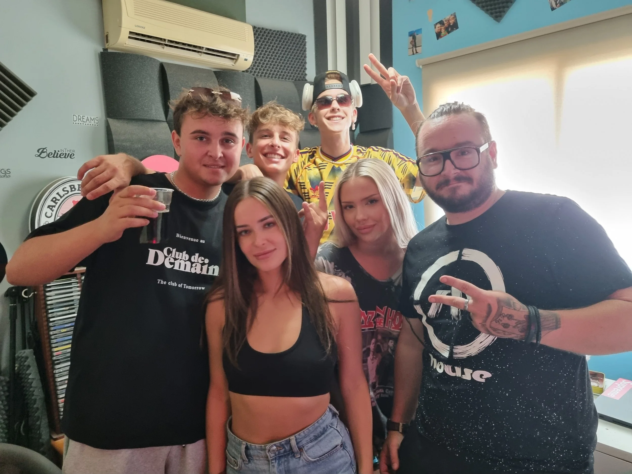 Group of six young people posing for a photo in a music studio. Some are holding drinks, and one person is wearing headphones and sunglasses. They are smiling and making peace signs. The background shows soundproofing panels, a window, and various po