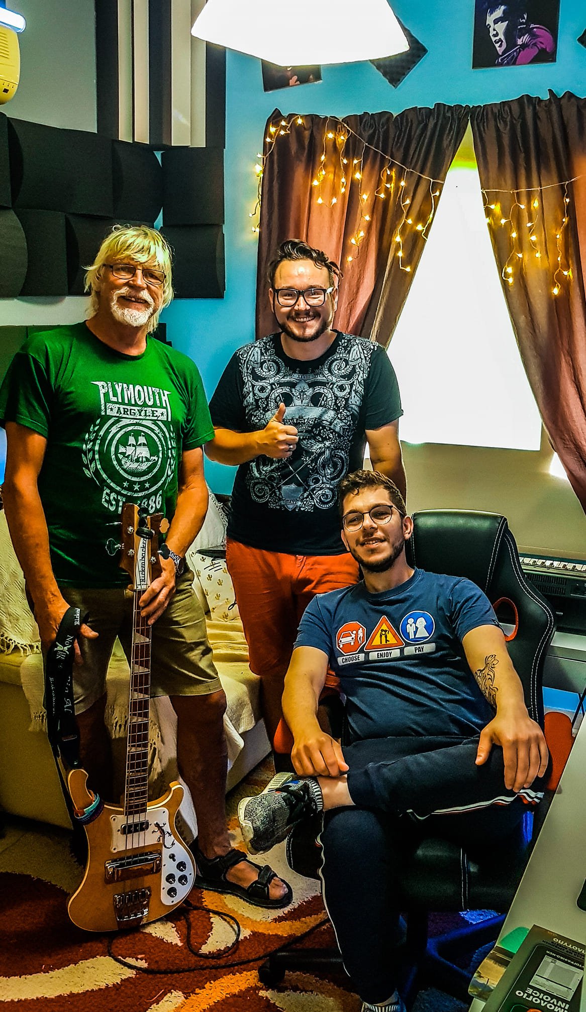 Four men smiling in a room with acoustic foam panels and string lights; one seated at a desk, three standing, one holding a guitar, one giving a thumbs up, a window with curtains behind them.