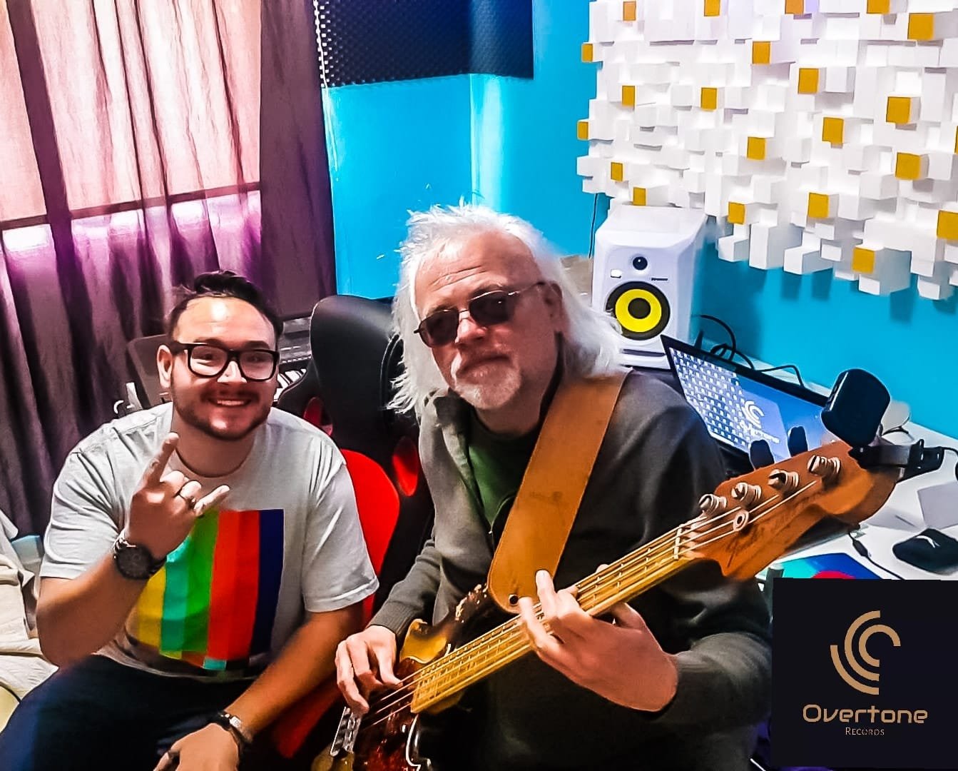 Two men in a recording studio, one with glasses and a rainbow t-shirt, making a rock sign, and the other older with gray hair, sunglasses, and a guitar. Studio equipment is visible in the background, including a speaker and acoustic wall panels.