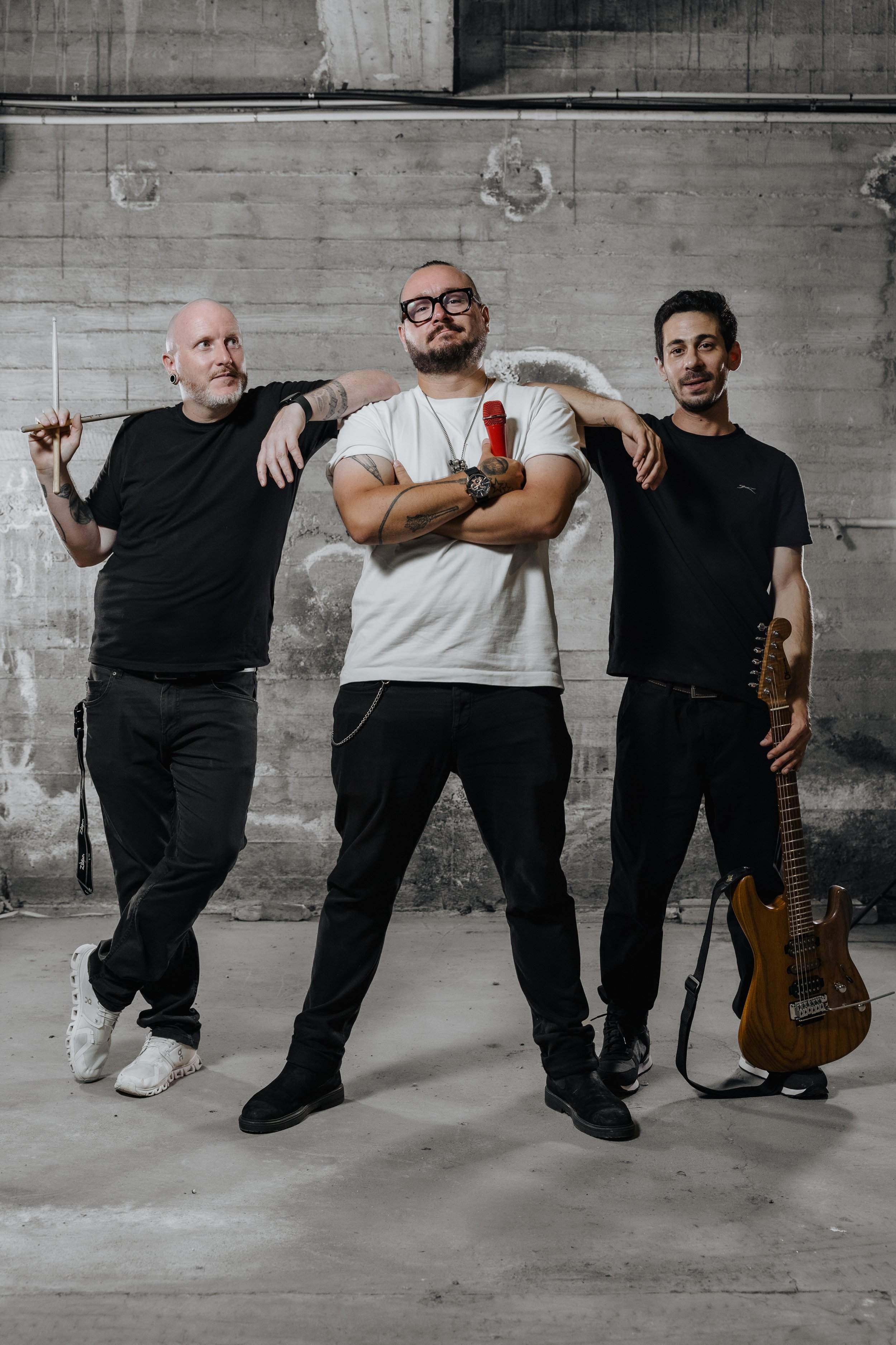 Three men standing in front of a concrete wall. The man in the middle has a red microphone, glasses, and tattoos, with arms crossed. The man on the left has behind him an arm on the middle man's shoulder and is holding a stick with a paddle. The man on the right holds an electric bass guitar. All are dressed casually in black or white.