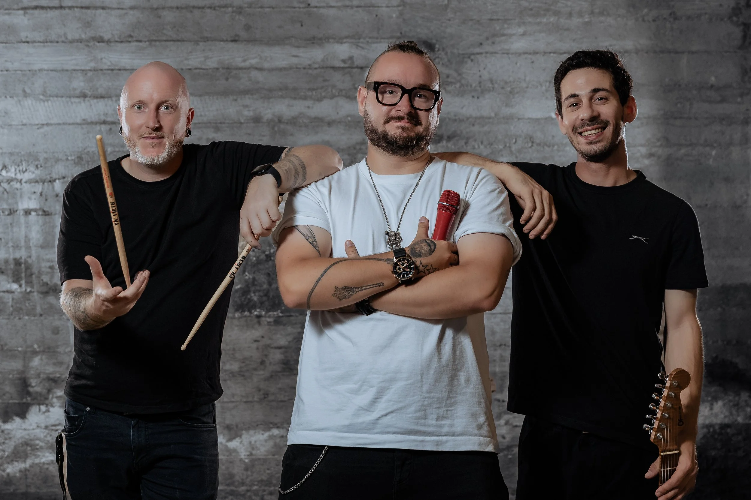Three men standing together against a gray wooden wall, smiling. The man in the middle is holding a red microphone, wearing glasses, a white t-shirt, and has tattoos on his arms. The man on the left has a shaved head, earrings, and is holding drumsti