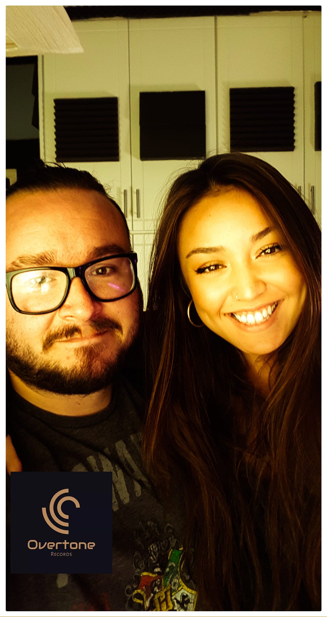 A man and woman smiling together, with a background of soundproof panels and white cabinets, and a logo for OverTone Records in the lower left corner.