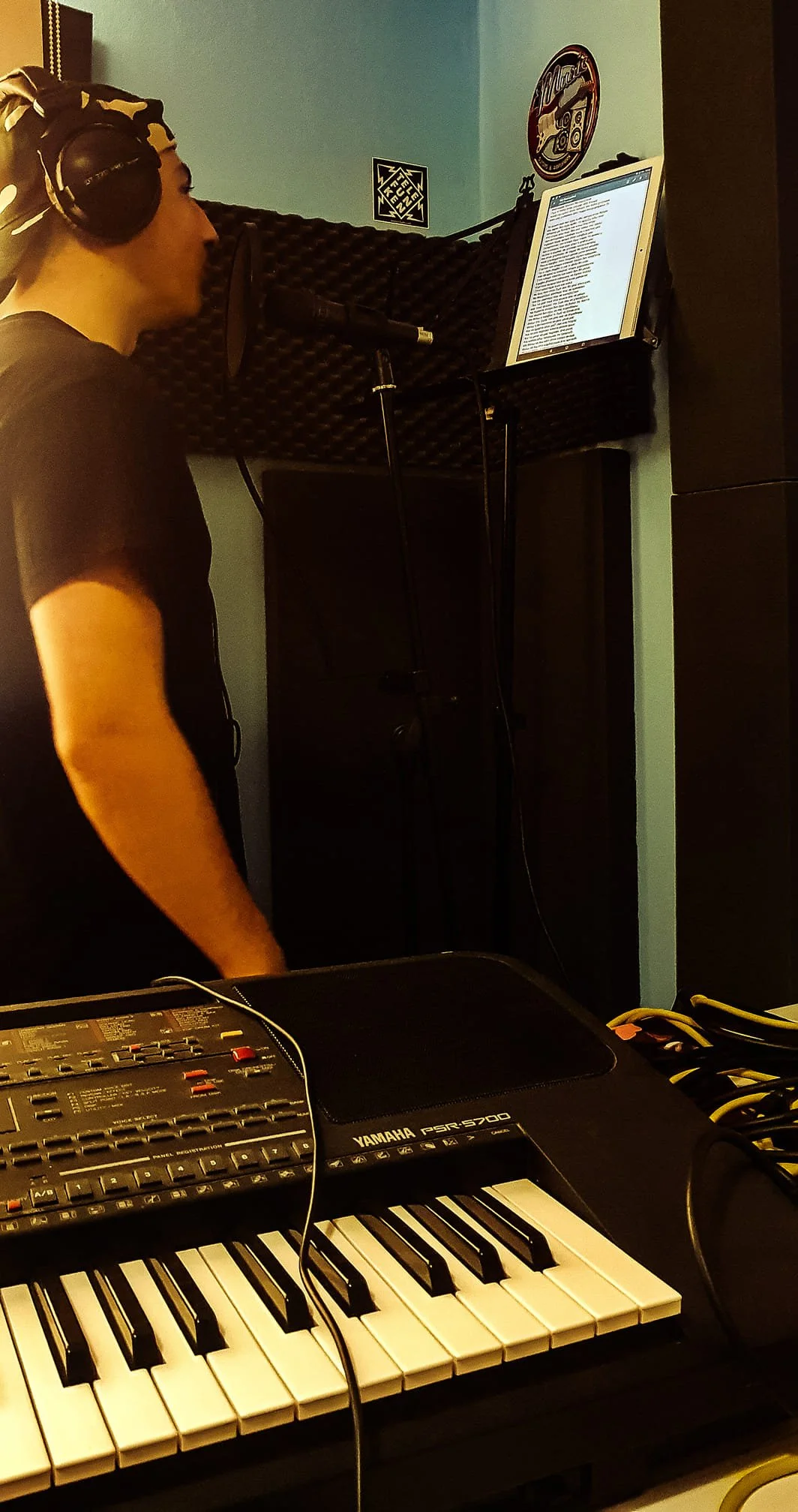 A person in a recording studio stands at a microphone with a tablet in front of them, surrounded by music equipment including a Yamaha keyboard, a mixer, and studio soundproofing panels on the wall.