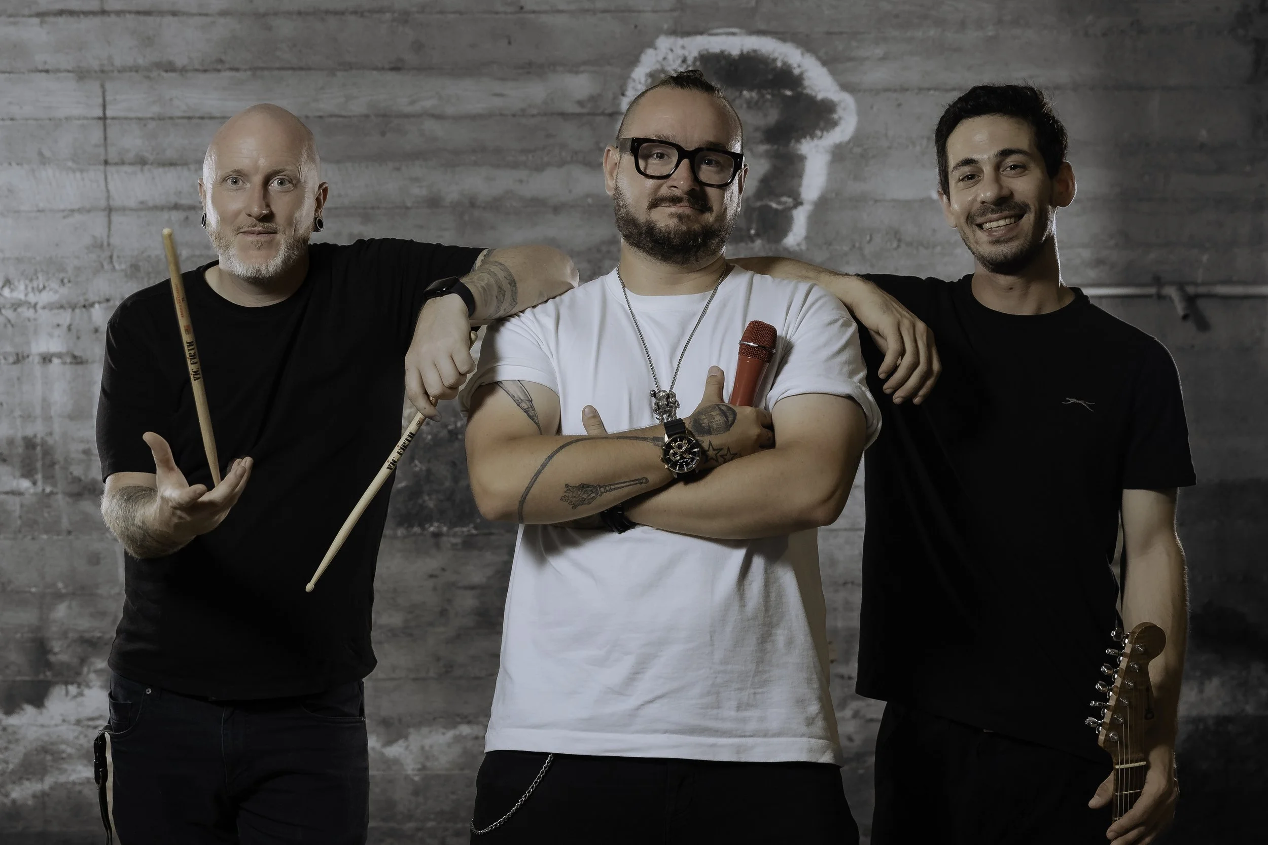 Three men standing together in front of a gray wooden wall. The man on the left holds drumsticks, the man in the middle holds a red microphone and has tattoos, glasses, and a beard, and the man on the right is holding a guitar headstock, smiling.