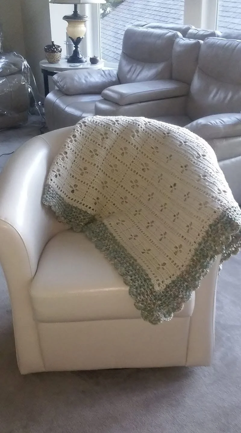 Cream-colored armchair with a crocheted blanket draped over the back in a living room with a leather sofa and a side table with lamp.