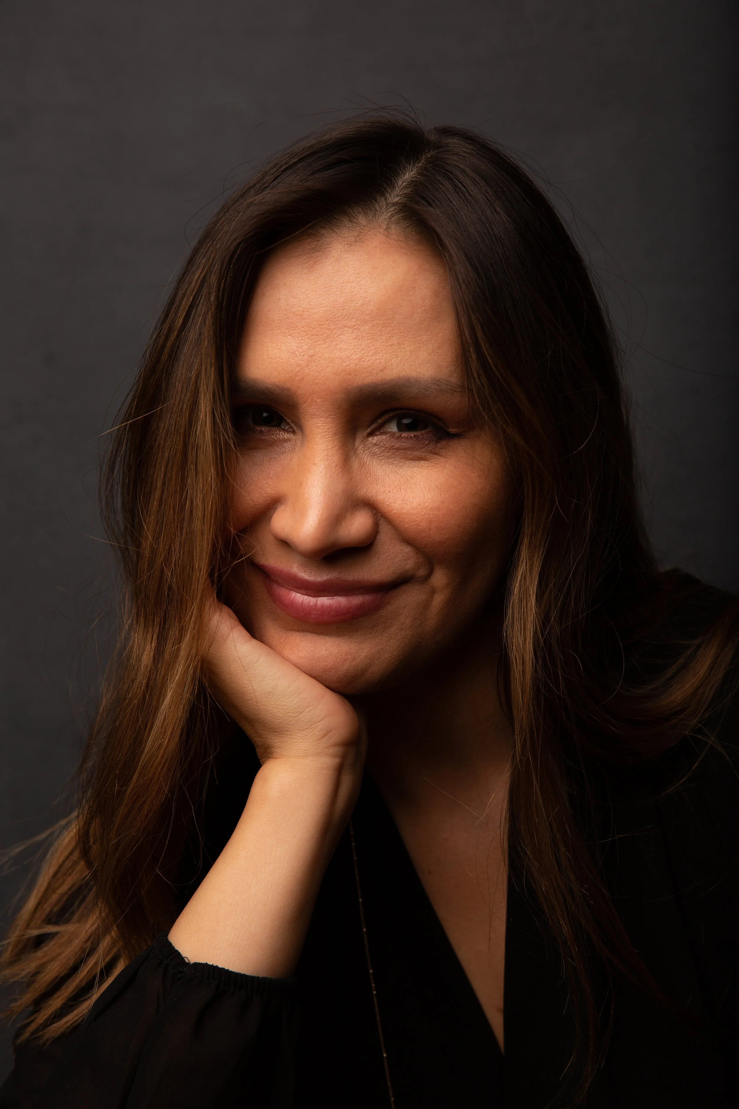 Close-up portrait of a woman with long brown hair, smiling, with her hand resting on her chin, against a dark background.