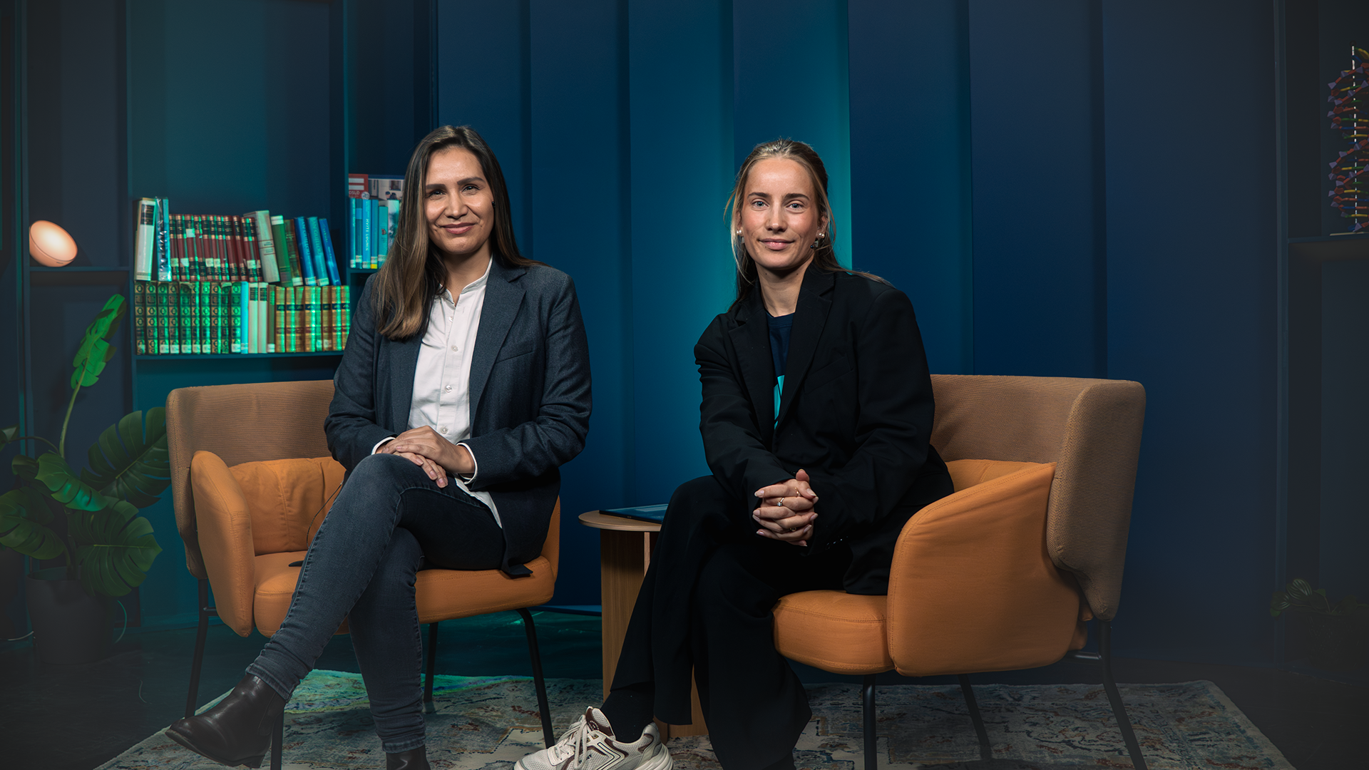 Two women sitting on orange chairs in a room with blue walls, bookshelves, and plants, looking at the camera.