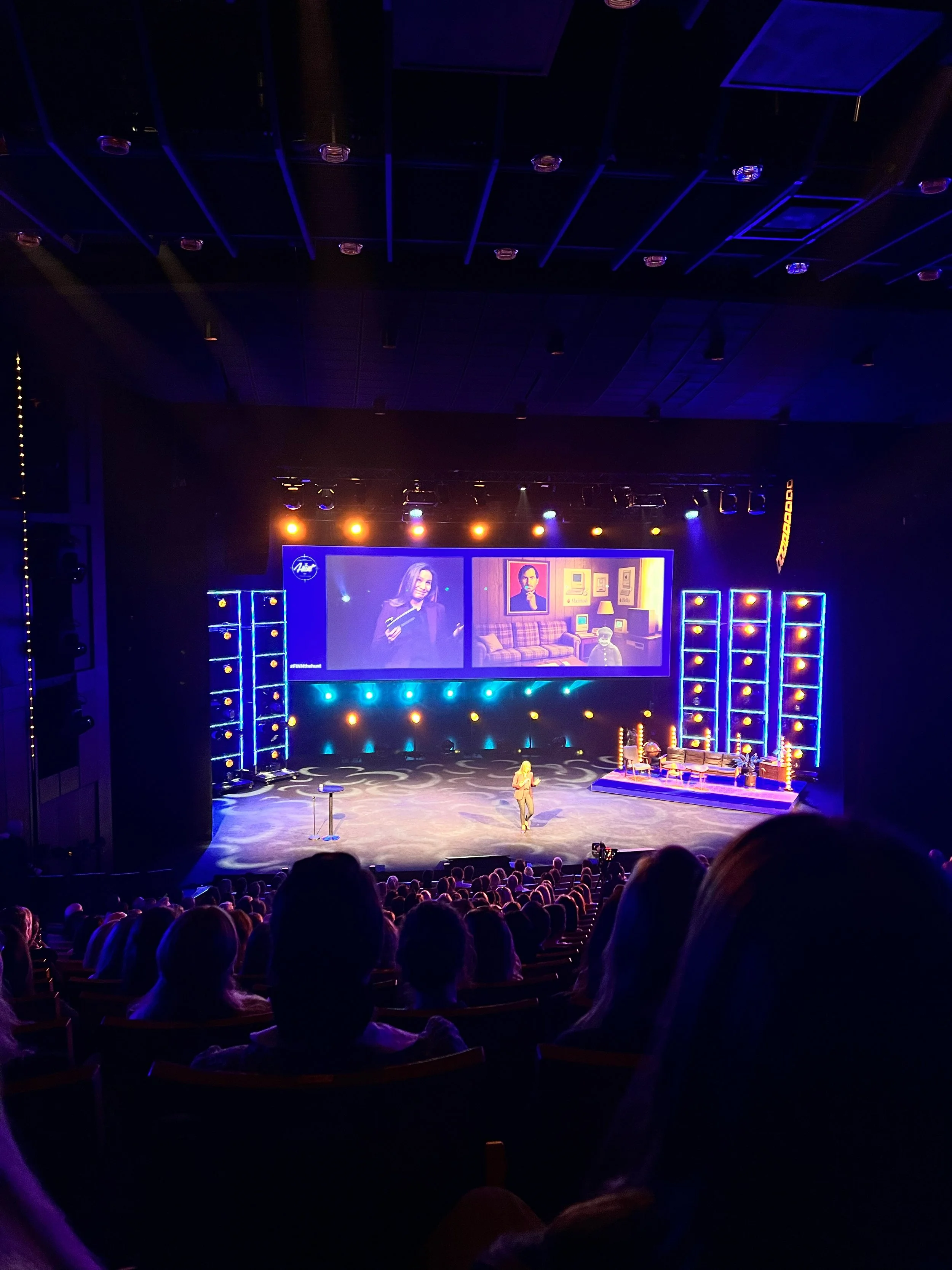 A stage with a speaker in front of a seated audience. The stage has large screens displaying a woman speaking and a man with items on a shelf behind him. The stage is lit with blue and yellow lights, and there are chairs and lighting equipment visible.
