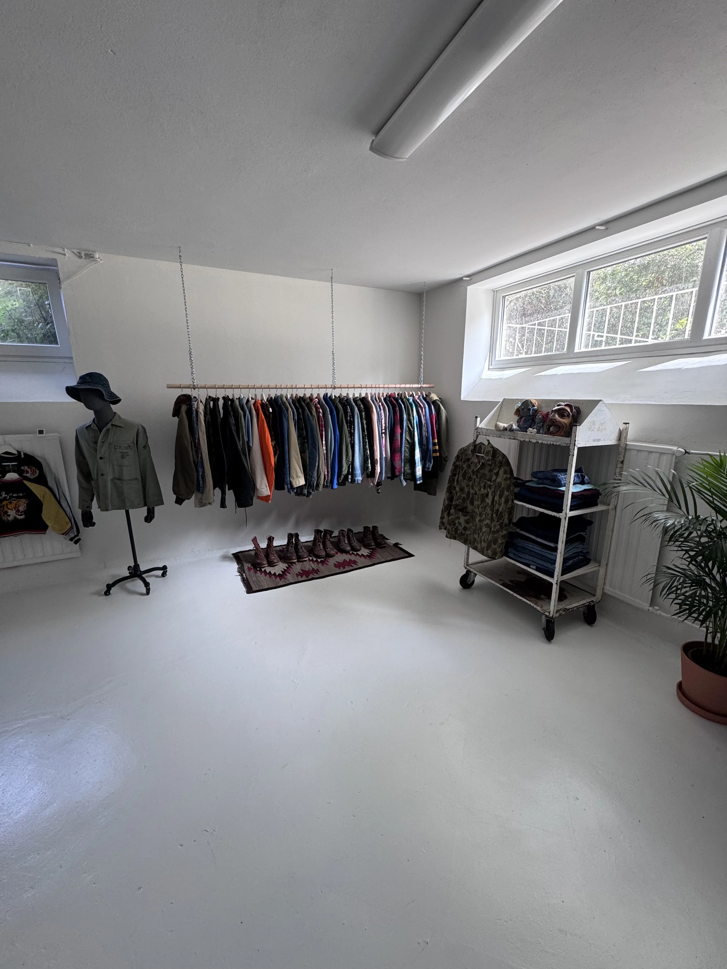 Interior of a clothing boutique or thrift store with a white floor, white walls, and a large window letting in natural light. There is a clothing rack with various jackets and shirts, a mannequin wearing a hat and jacket, a small cart filled with home decor or masks, and a potted plant in the corner.