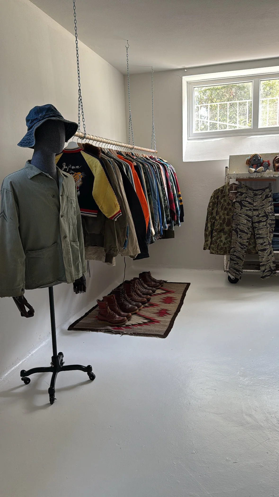 Indoor clothing store with a mannequin wearing a green jacket and a blue hat, a clothing rack with various jackets and shirts, a small rug with boots lined up, a window letting in natural light, and a display with stuffed animals.