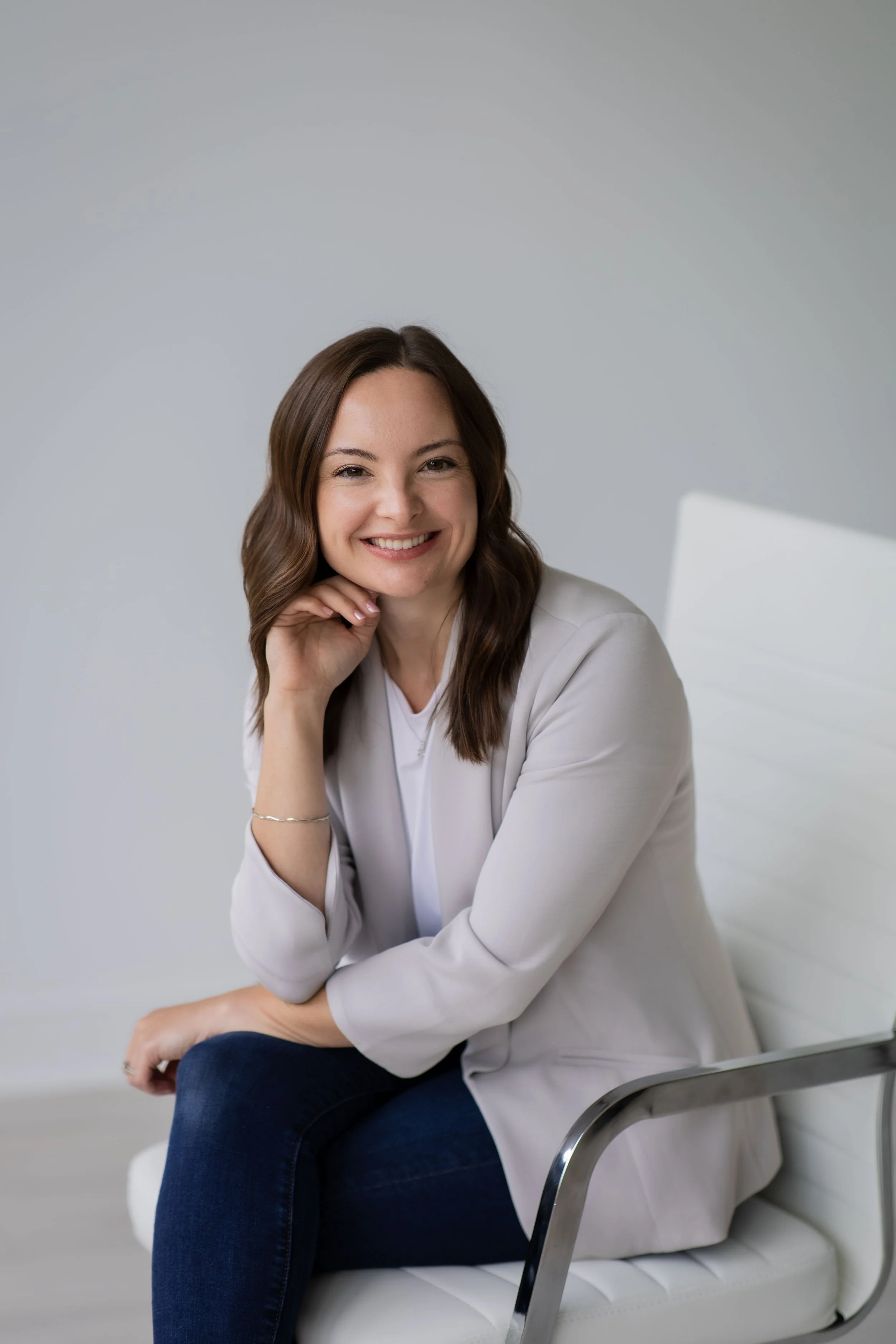 A smiling woman with brown hair sitting on a white bench in a bright room with a gray background. She is wearing a light gray blazer, white top, and dark blue jeans, resting her chin on her hand and looking at the camera.