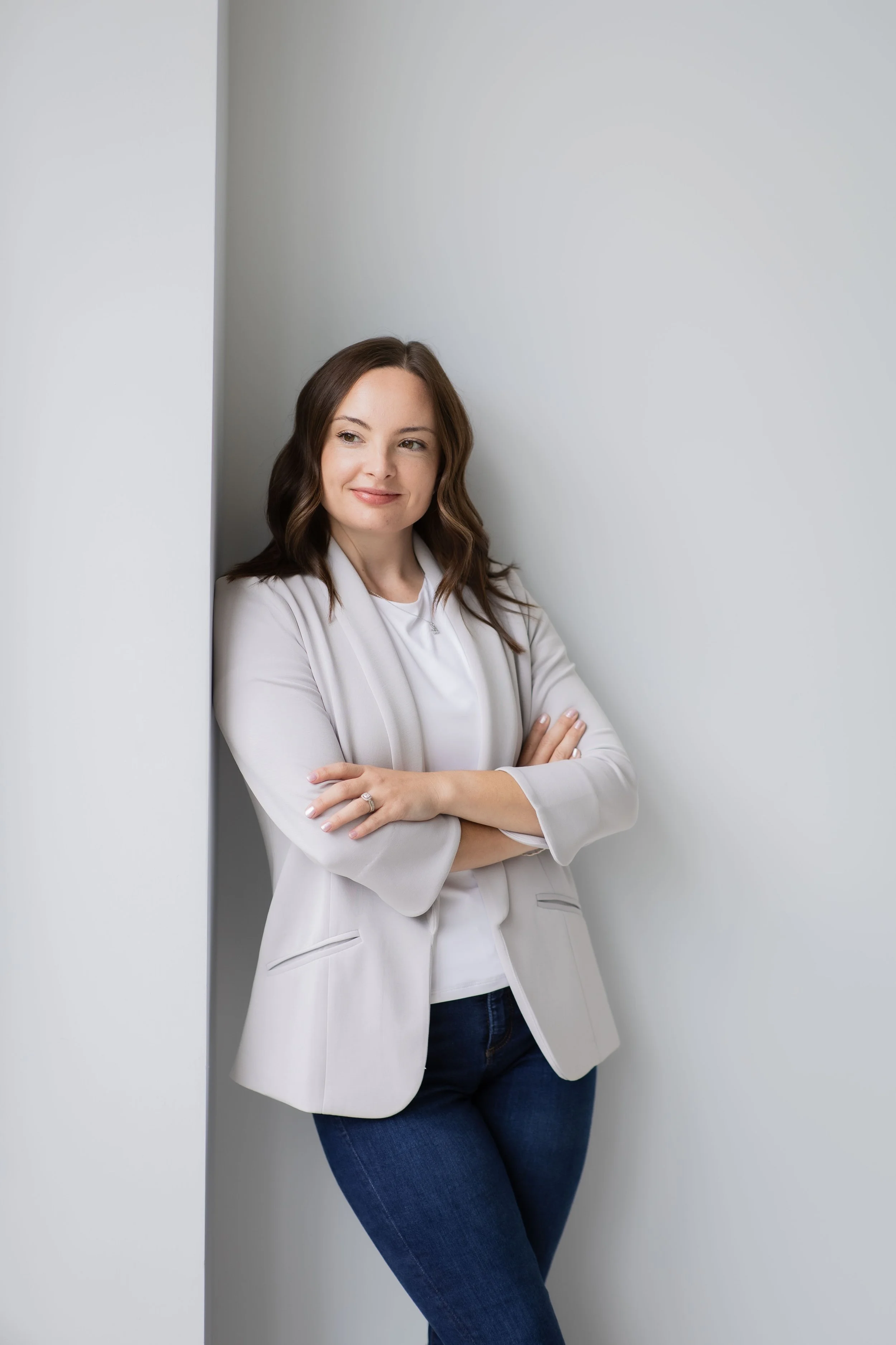 Woman with shoulder-length brown hair, wearing a white blazer and blue jeans, standing with her arms crossed and leaning against a wall with a slight smile.