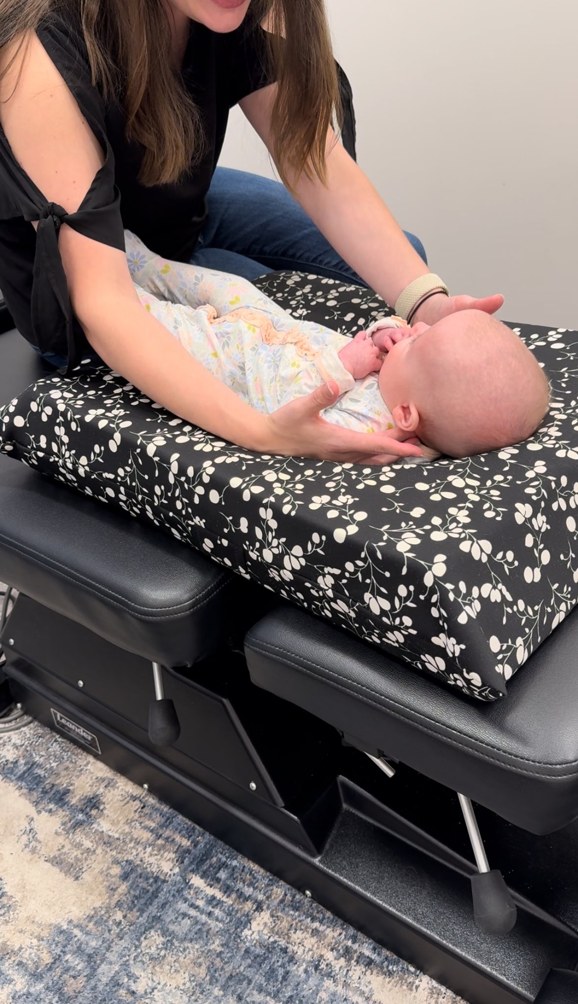 A woman performing a medical examination or chiropractic adjustment on a baby lying on an examination table with a black and white floral patterned surface.