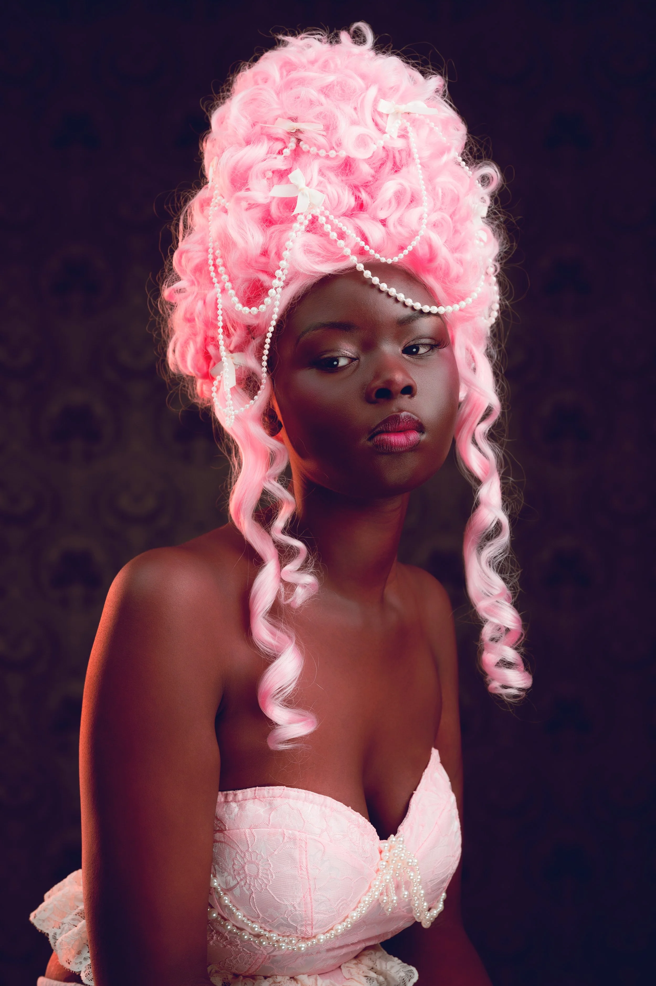 A woman with dark skin and pink, curly hair styled in an elaborate updo adorned with pearl jewelry and bows, wearing a strapless pink lace dress, standing against a dark, patterned background.