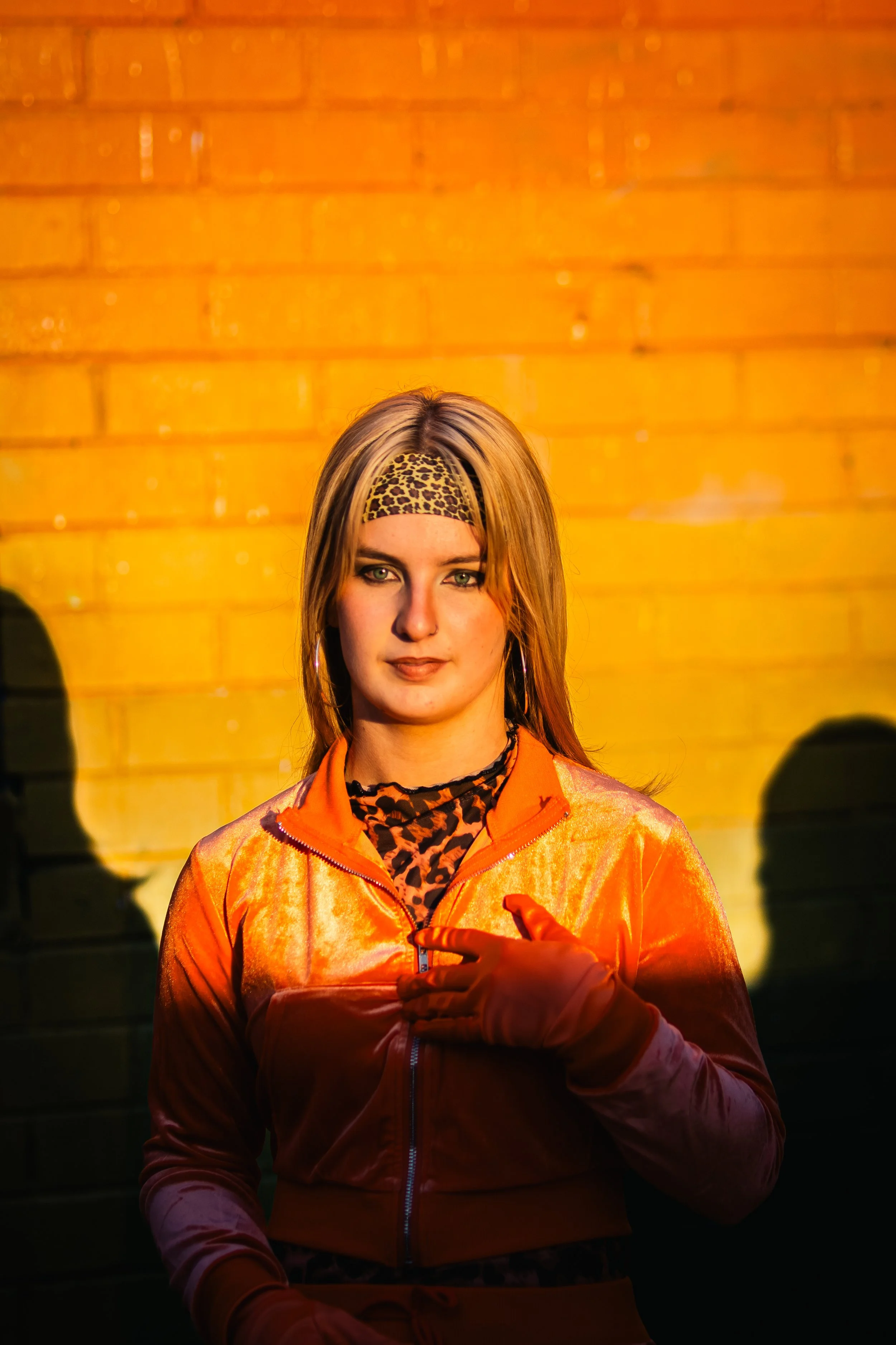 A young woman with blonde hair and green eyes standing against a yellow brick wall, wearing a leopard print headband, orange jacket, and zebra print shirt, with shadows cast on the wall behind her.