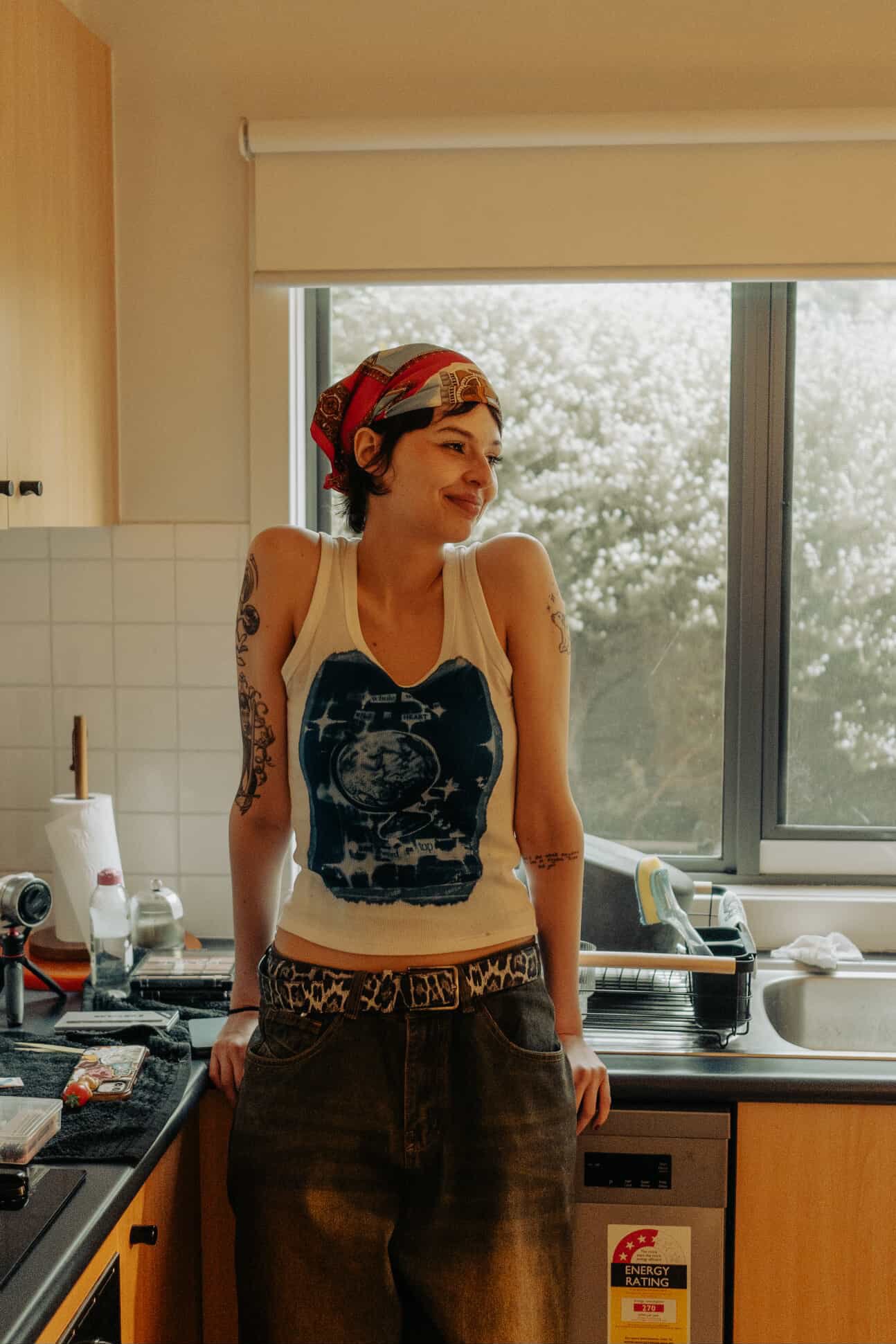 A woman with short dark hair wearing a bandana, tank top, and dark pants standing in a kitchen next to a window with a view of trees outside.