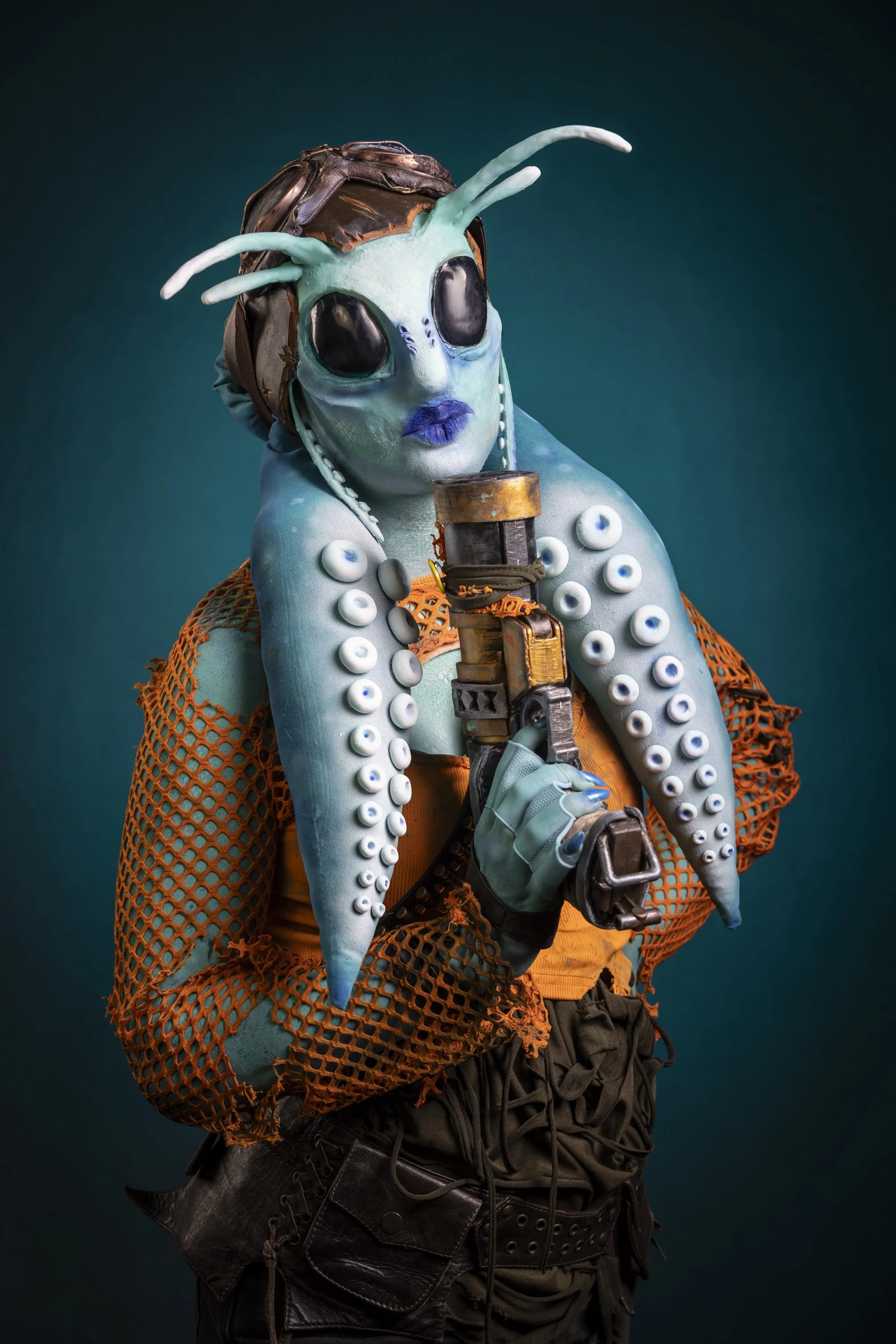 A person wearing alien and sci-fi inspired costume and accessories, including a large alien mask with antennae, a textured costume with bubble-like details, and holding a futuristic prop weapon against a dark teal background.