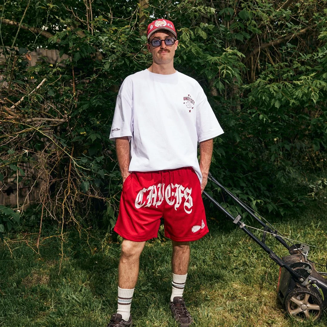 A man standing outside in a backyard wearing a white oversized T-shirt, red shorts with 'CHAOS' written on them, striped socks, sneakers, sunglasses, and a red cap. He is holding a lawn mower.