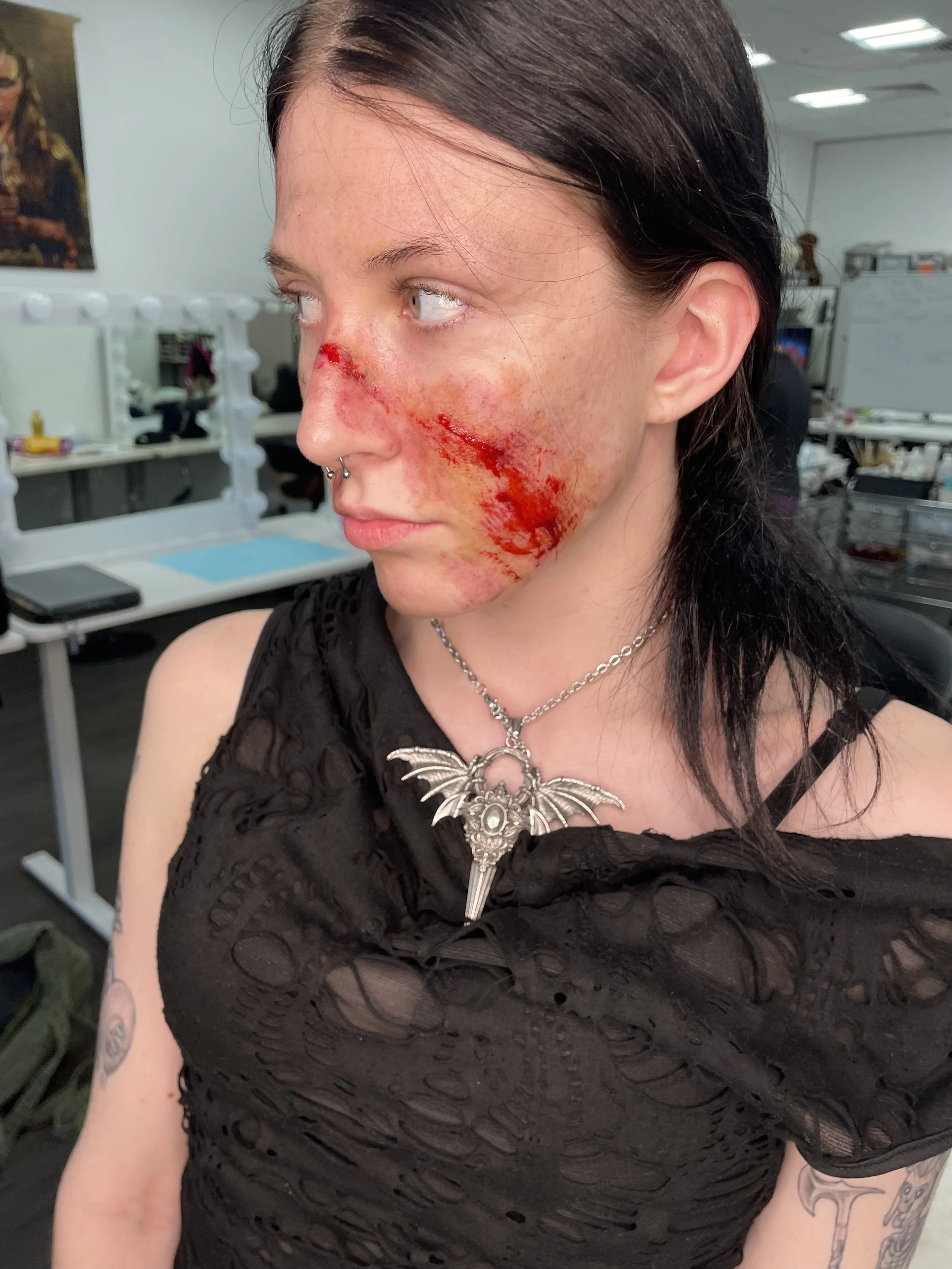 A woman with dark hair and nose piercings has a large, fresh facial wound with blood running down her cheek, in an indoor office or studio setting.