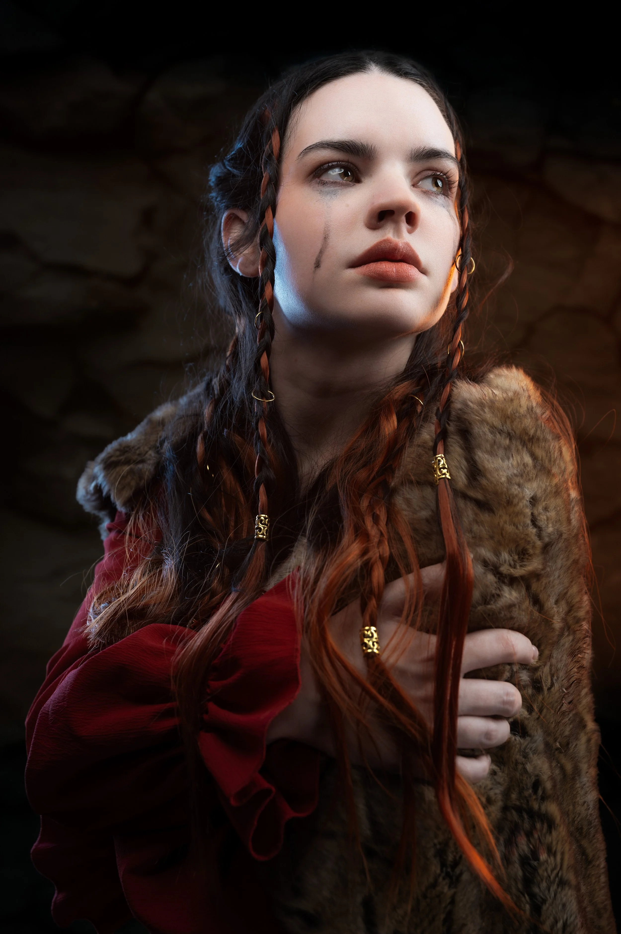 A young woman with long, curly brown hair and braided strands, wearing a red dress with ruffled sleeves and a fur stole, holding her hand close to her chest, against a dark, stone wall background.