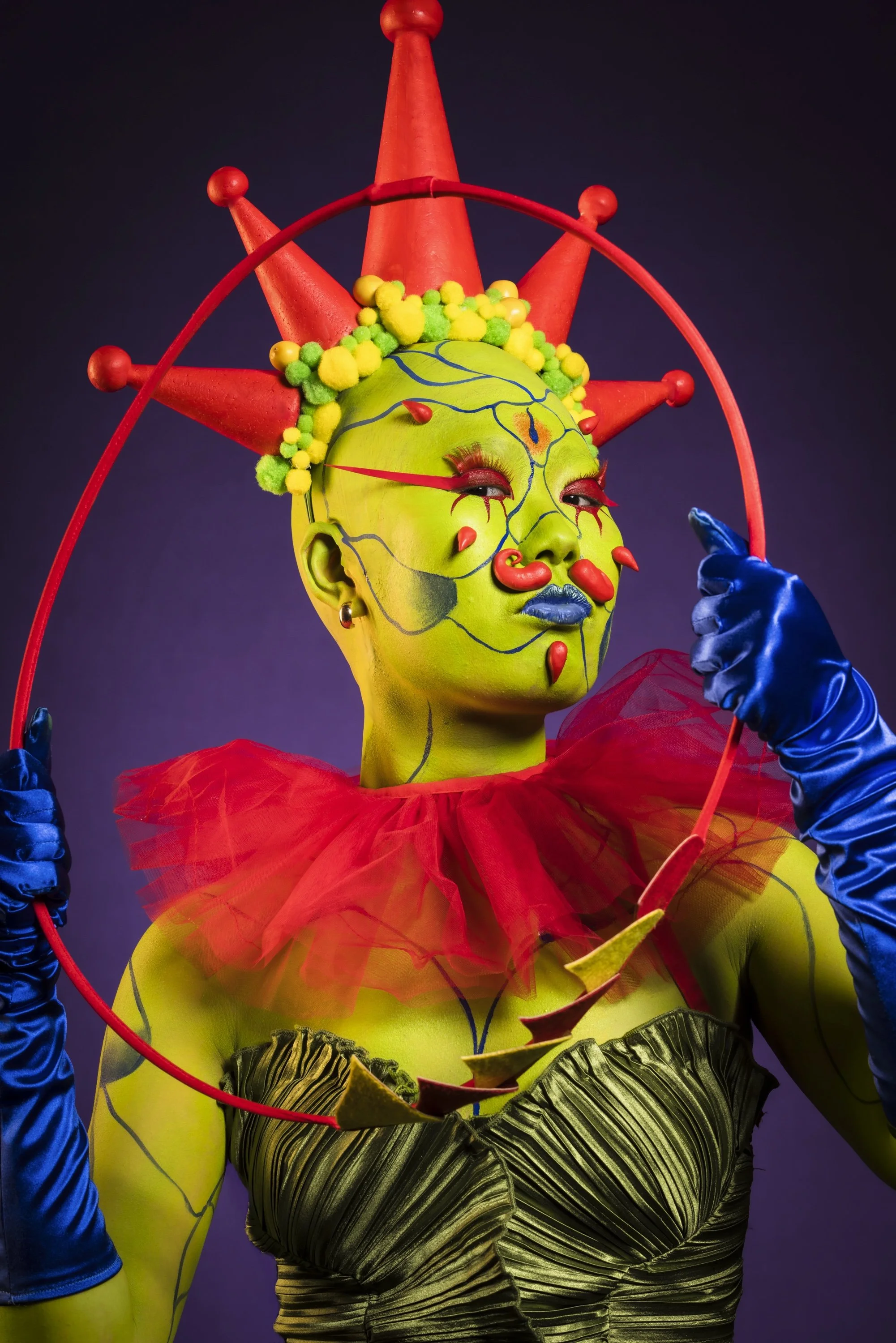 A person with bright green body paint, stylized makeup, and elaborate costume, including a headpiece with red cones and colorful decorations, holding a red hoop against a dark background.