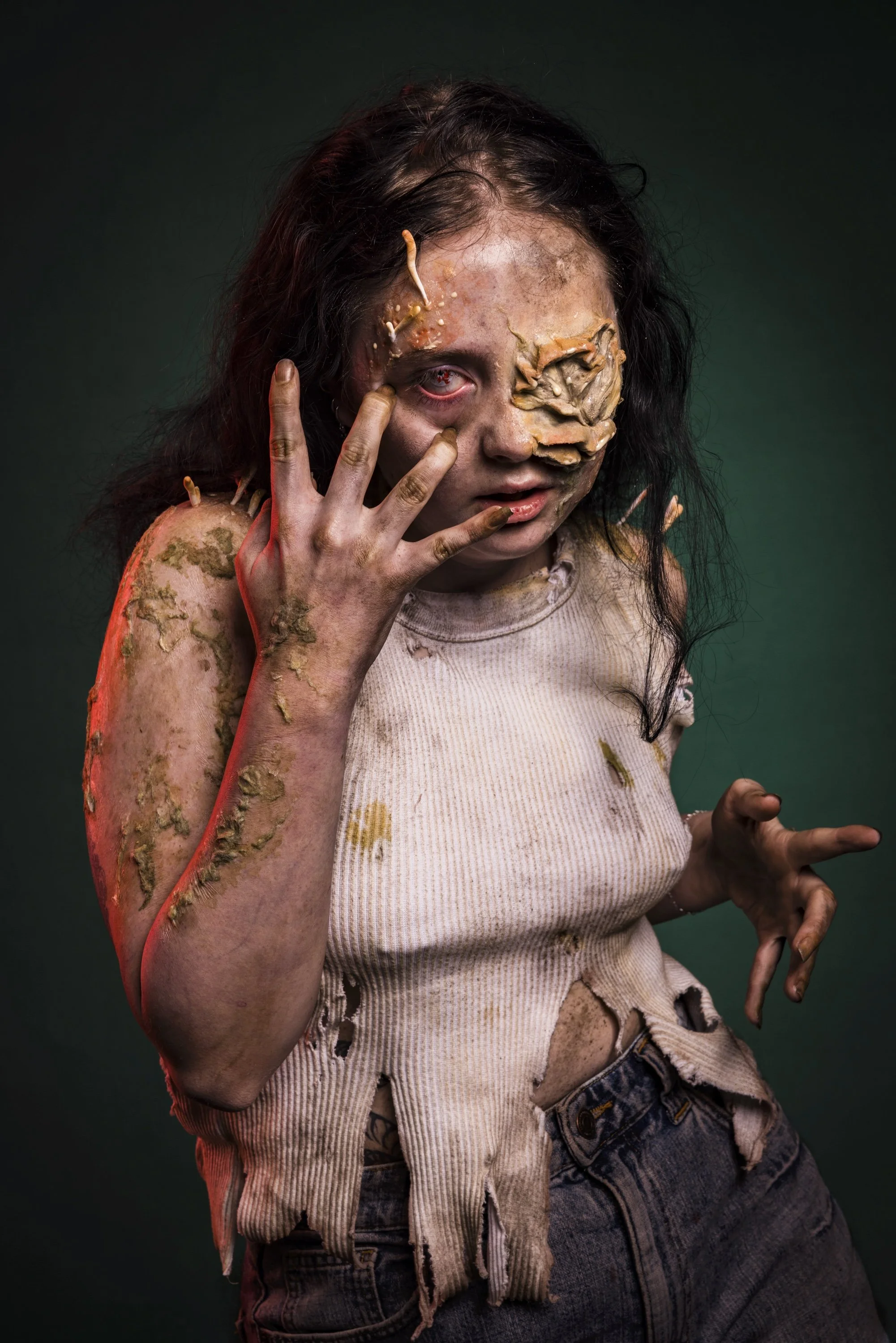 A person with disheveled dark hair and a distressed expression, covered in mud and deformities, wearing a torn white tank top and jeans, posing against a dark green background.