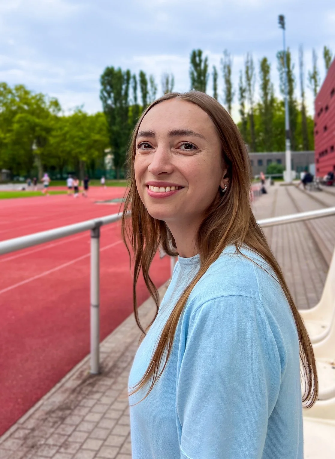 The photo shows the sports psychologist Eva Sippel smiling warmly. She has long brown hair, is wearing a light blue top and is standing at the edge of an athletics stadium in Berlin.