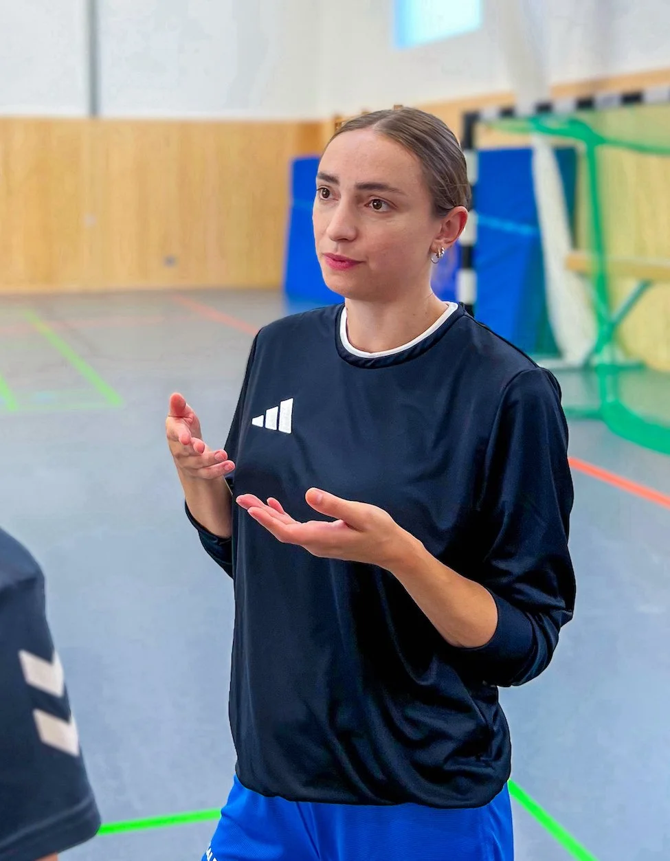 In this photo, the sports psychologist Eva Sippel is standing in a sports hall. She is talking to another person and looks somewhat serious, but engaged. She is wearing a dark Adidas sports top and blue tracksuit bottoms.