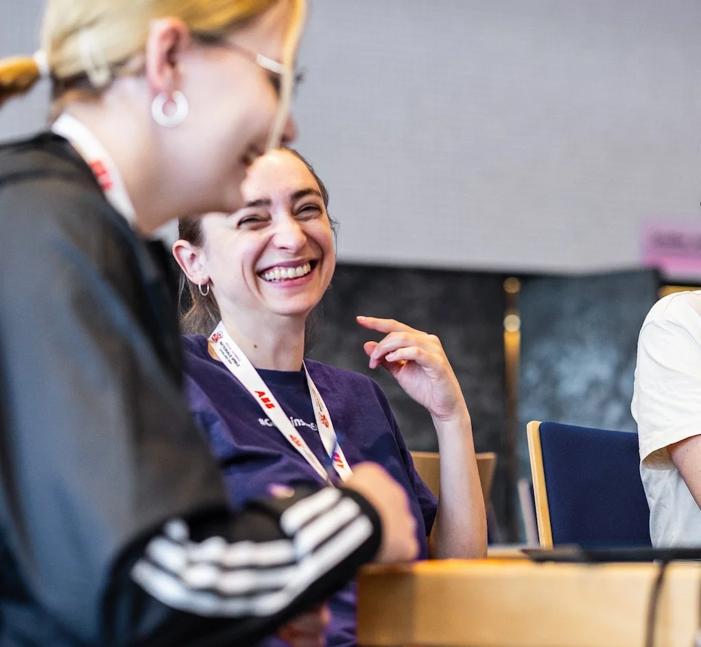 Zwei lachende Frauen im Gespräch bei einer Sportveranstaltung. Die Sportpsychologin Eva Sippel trägt ein Lanyard mit Akkreditierung.