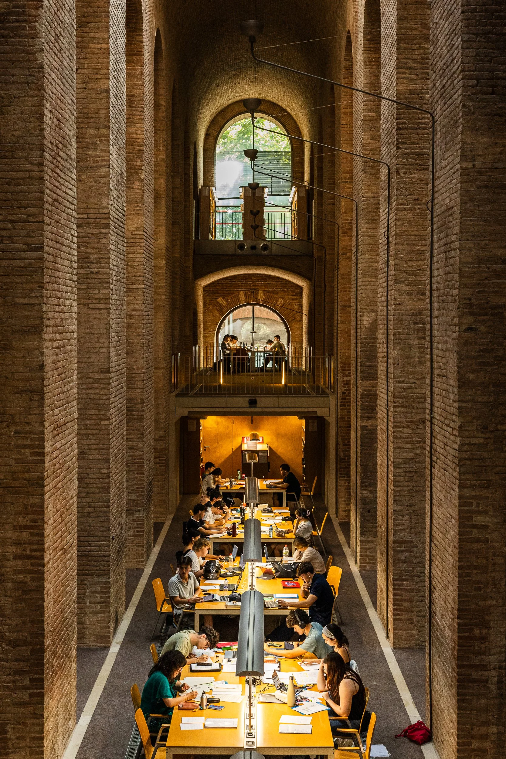 People working and studying at long tables in a large, high-ceilinged brick-walled library or study hall with natural light streaming through tall arched windows.