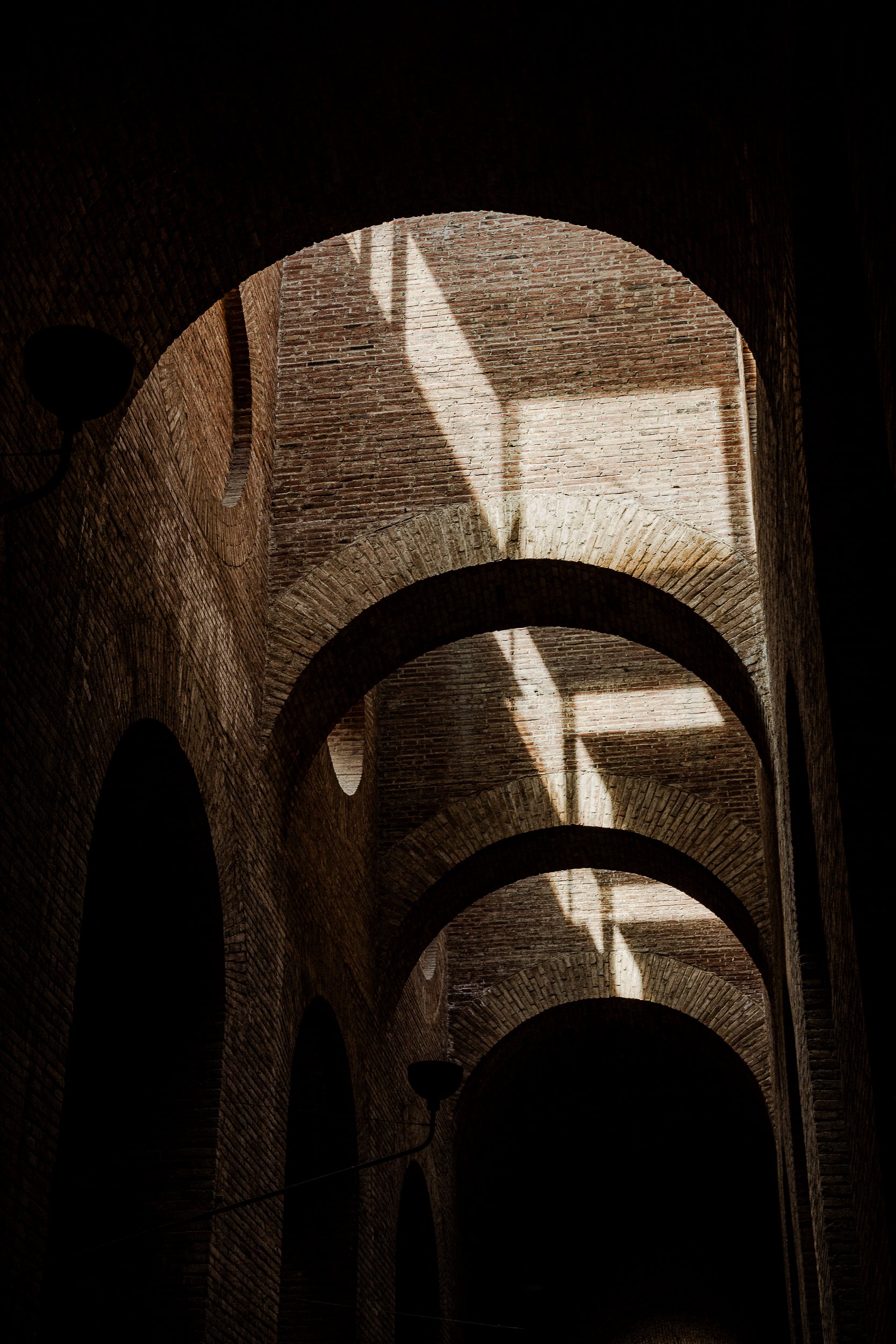 Repeating arches recede into darkness, a hard diagonal slash of light cutting across the brick.