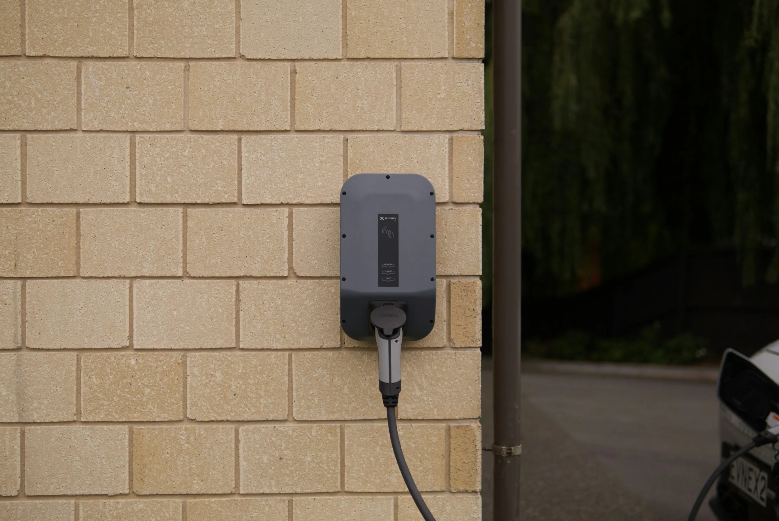 Electric vehicle charging station mounted on a beige brick wall with a cable plugged in, outdoors at dusk.