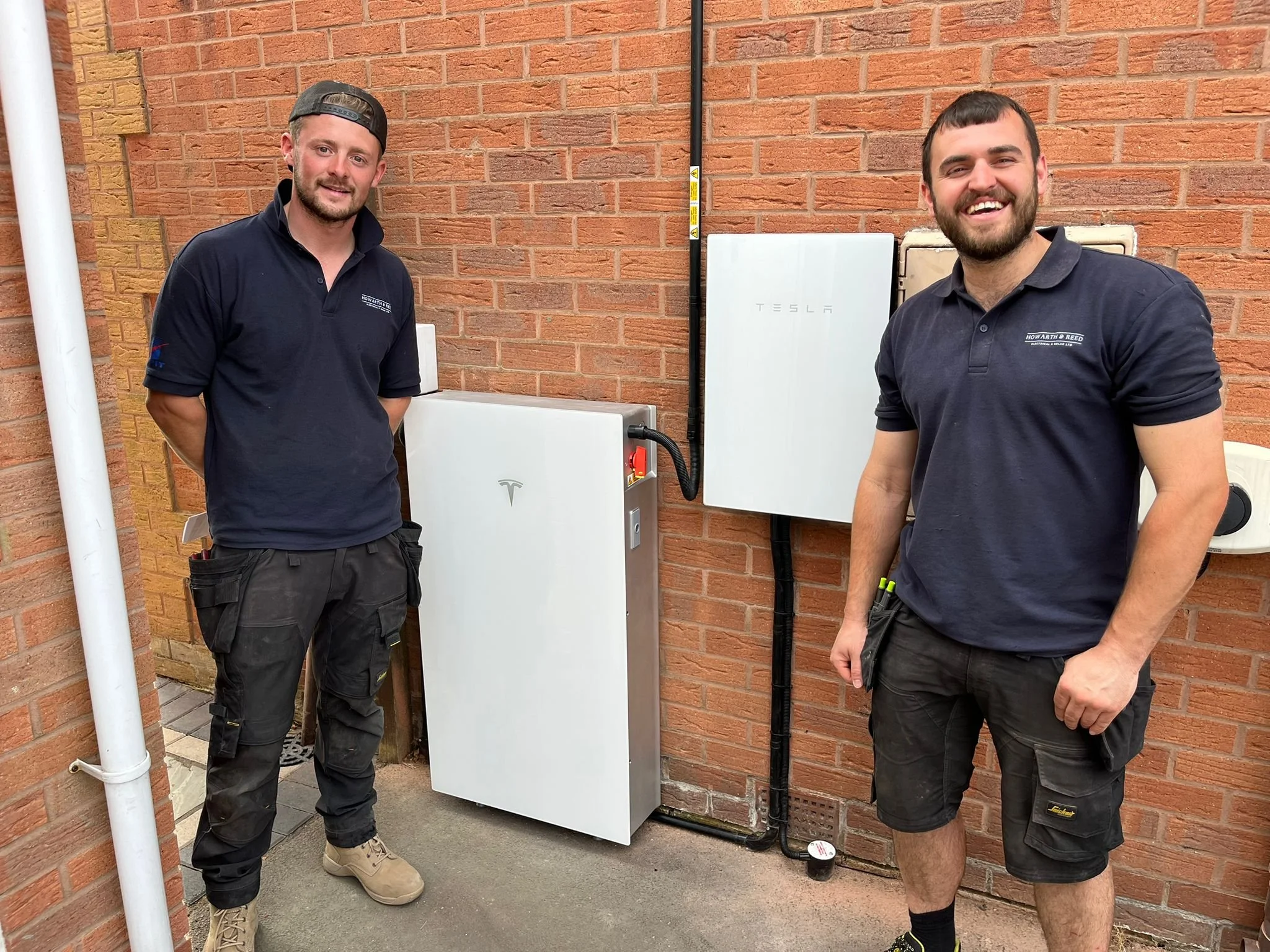 Two men standing outdoors near a brick wall, smiling, in front of Tesla electrical equipment mounted on the wall.