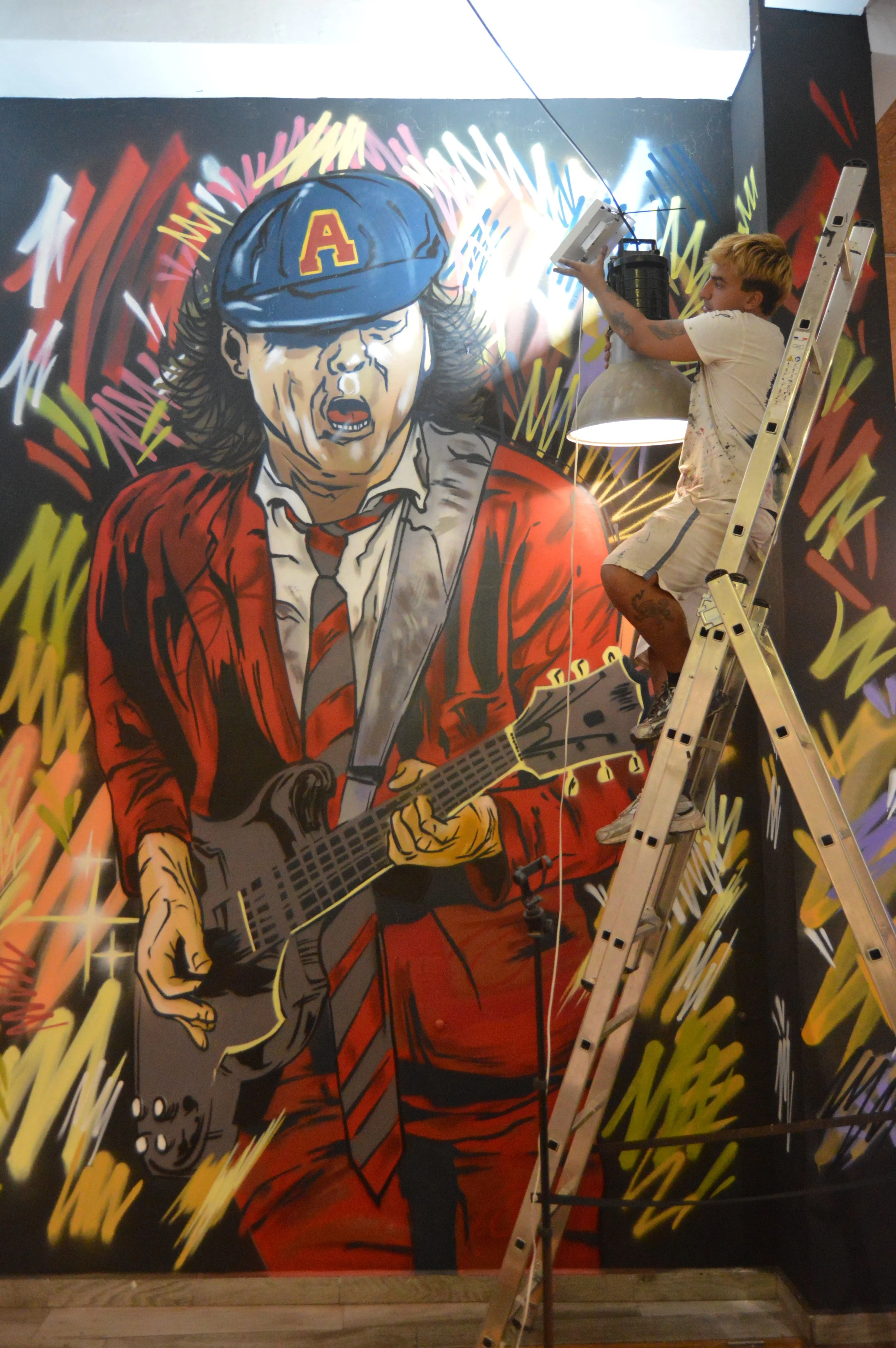 Un artista está pintando un mural estilo cómic de una mujer con un gorra azul con la letra 'A', vestida con saco rojo y camisa blanca, en una pared. Está en una escaleras y rodeado de colores vibrantes y rayos de luz en el fondo.