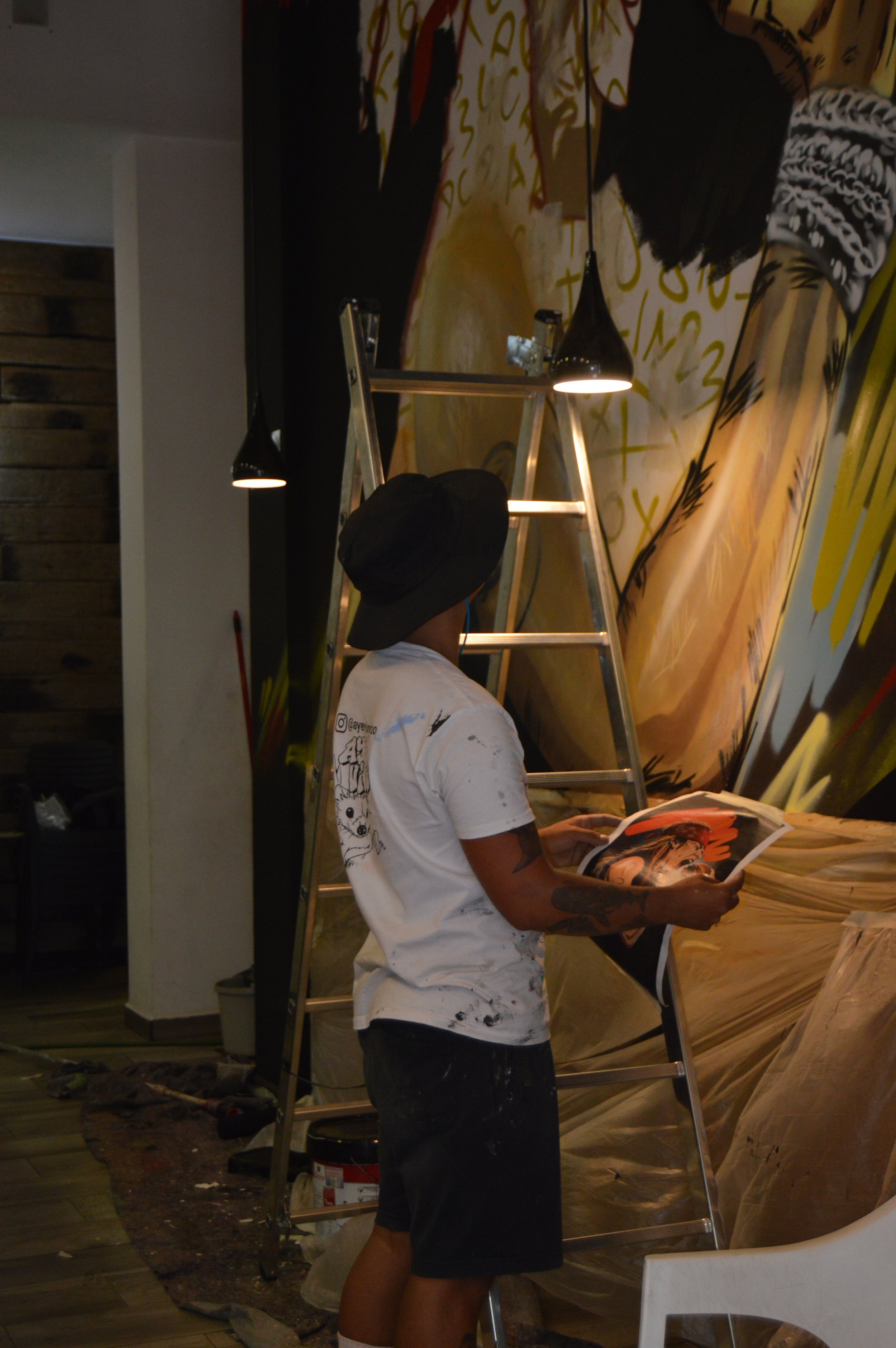 Persona de espaldas que lee una revista, de pie frente a una pared con pintura mural, usando sombrero negro, camiseta blanca con dibujos y pantalones cortos negros, cerca de una escalera de aluminio y luces de pared.