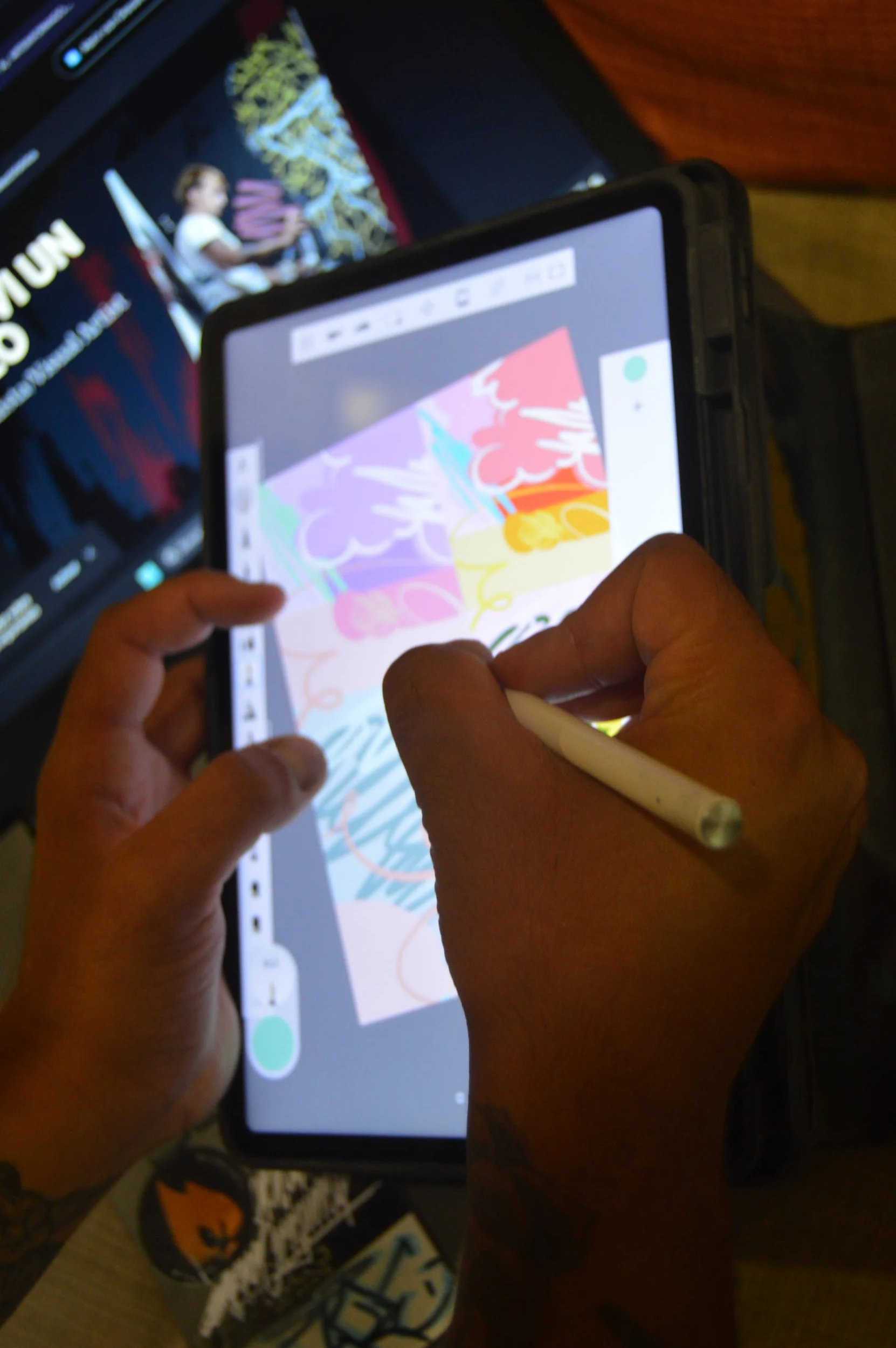 Persona dibujando en una tableta digital con un stylus, con otra pantalla visible en el fondo, en un entorno interior.