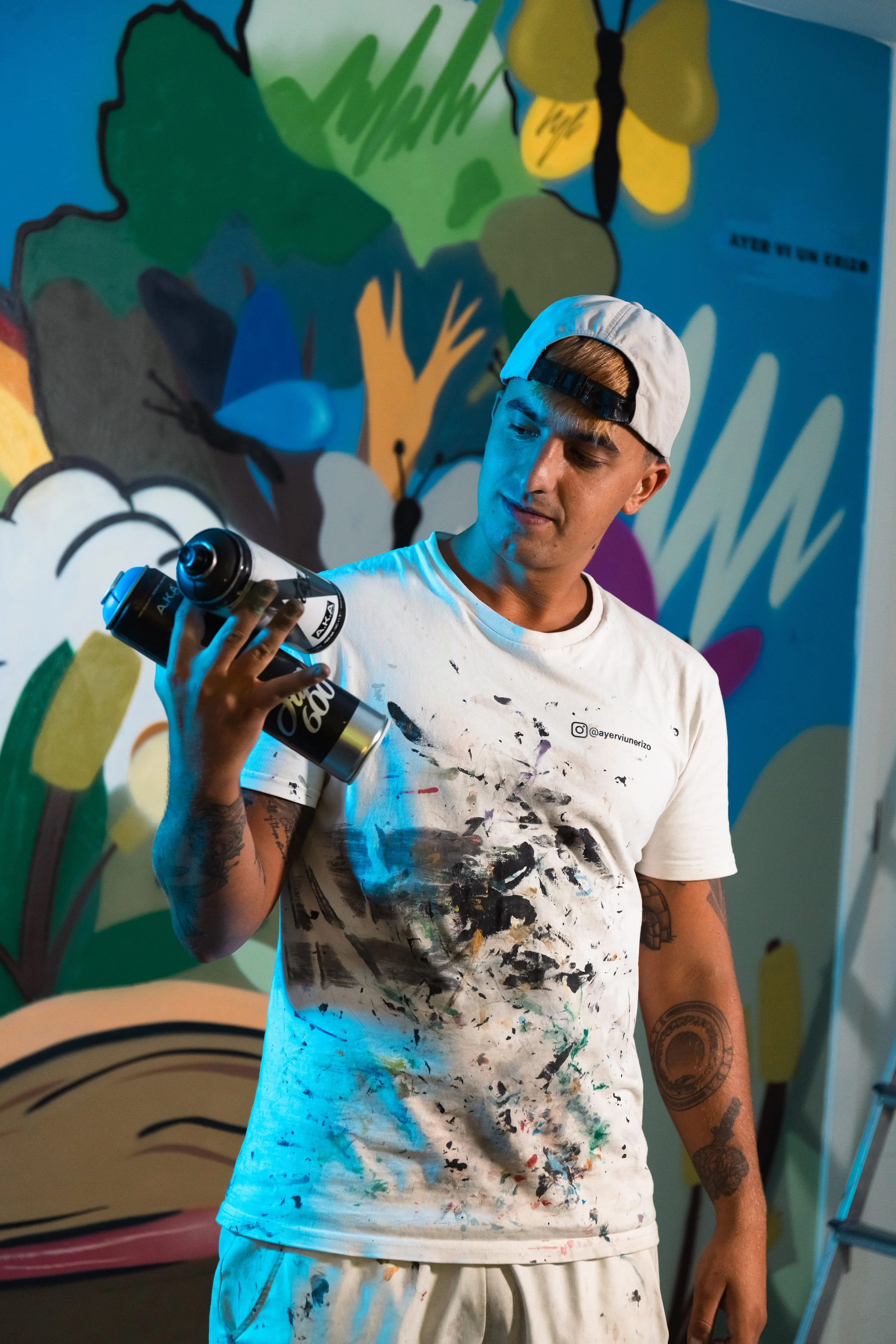 Un joven pintor con camiseta y pantalones sucios de pintura, sosteniendo una botella de agua mientras mira hacia abajo, en un fondo de mural colorido con temática de naturaleza y animales.