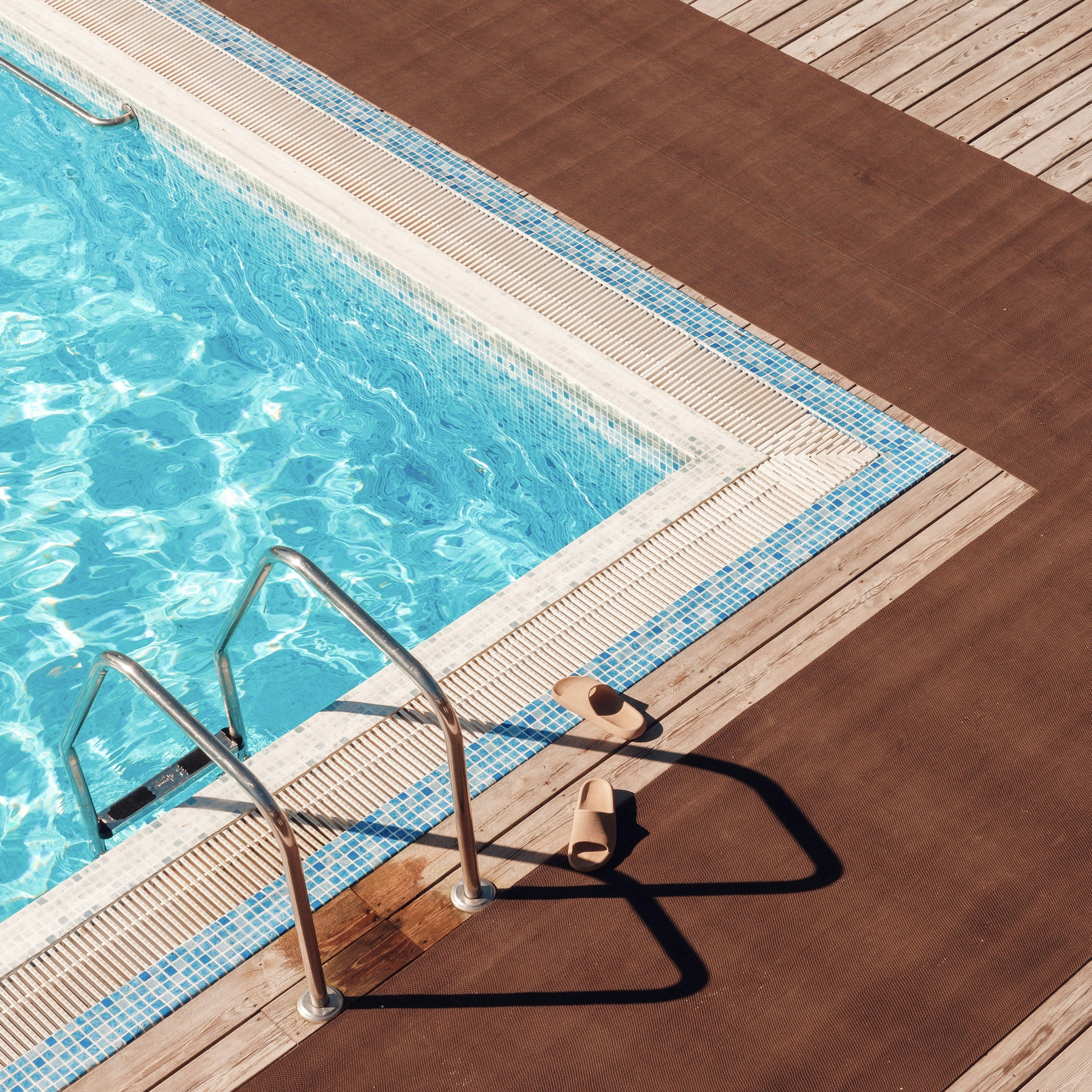 Swimming pool with clear blue water, pool stairs, and a brown wooden deck with a brown mat and a pair of sandals