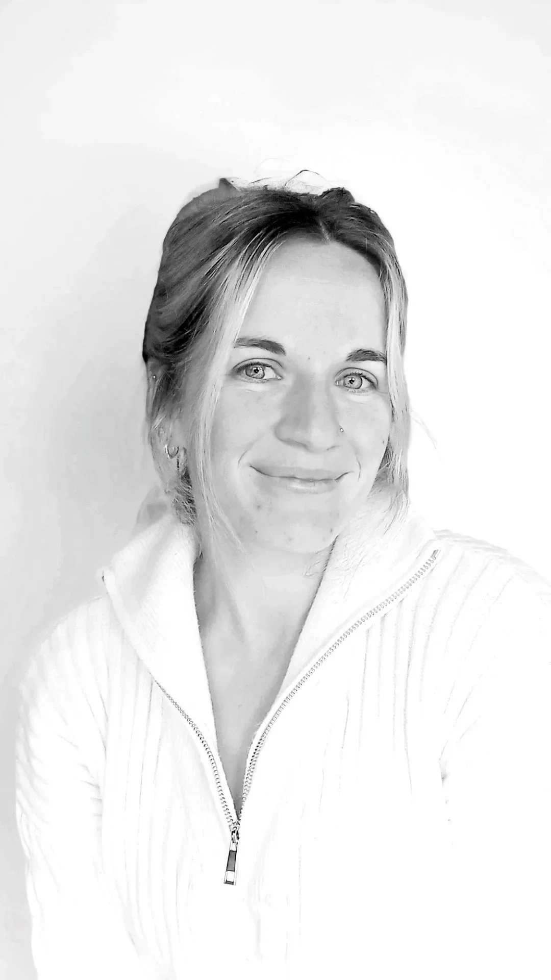 Black-and-white photo of a woman with shoulder-length hair, smiling, wearing a zip-up sweater standing against a plain background.