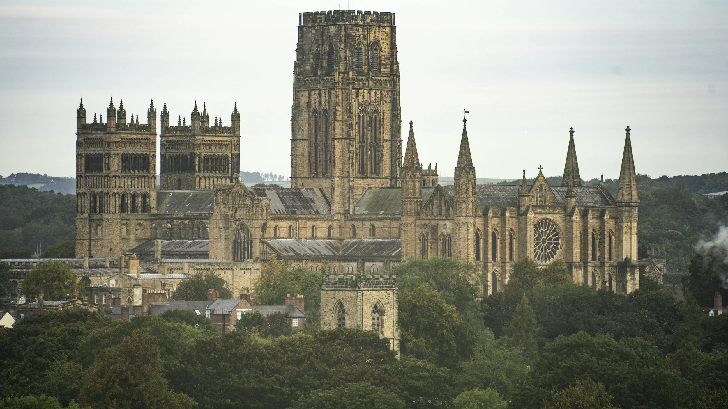 An old Gothic-style stone cathedral with tall spires and a large rose window, situated on a hill surrounded by trees and a small town at the base.