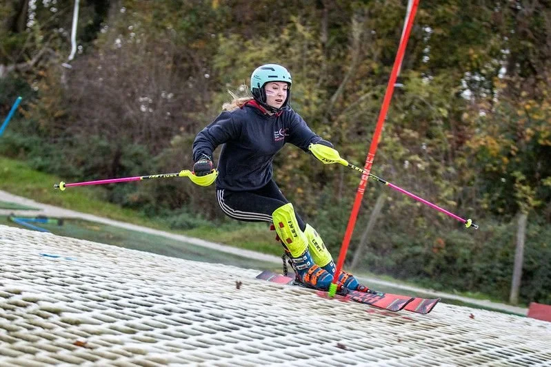 A woman wearing a helmet and protective gear skiing on a sloped surface outdoors, possibly a dry ski slope with trees in the background.