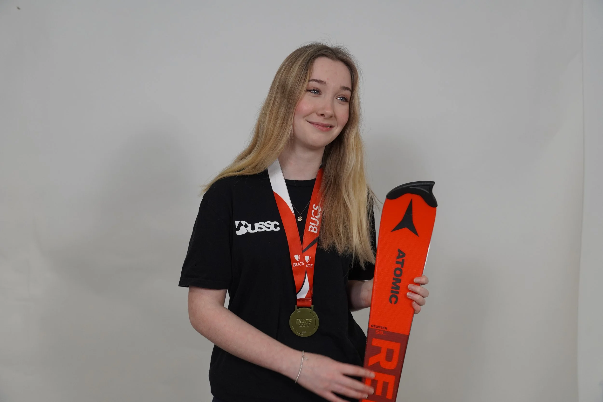 A young woman with long blonde hair and a black shirt is smiling slightly and holding a bright orange snowboard. She is wearing a gold medal around her neck and a black lanyard with red accents and white text. The background is a plain light gray wall.