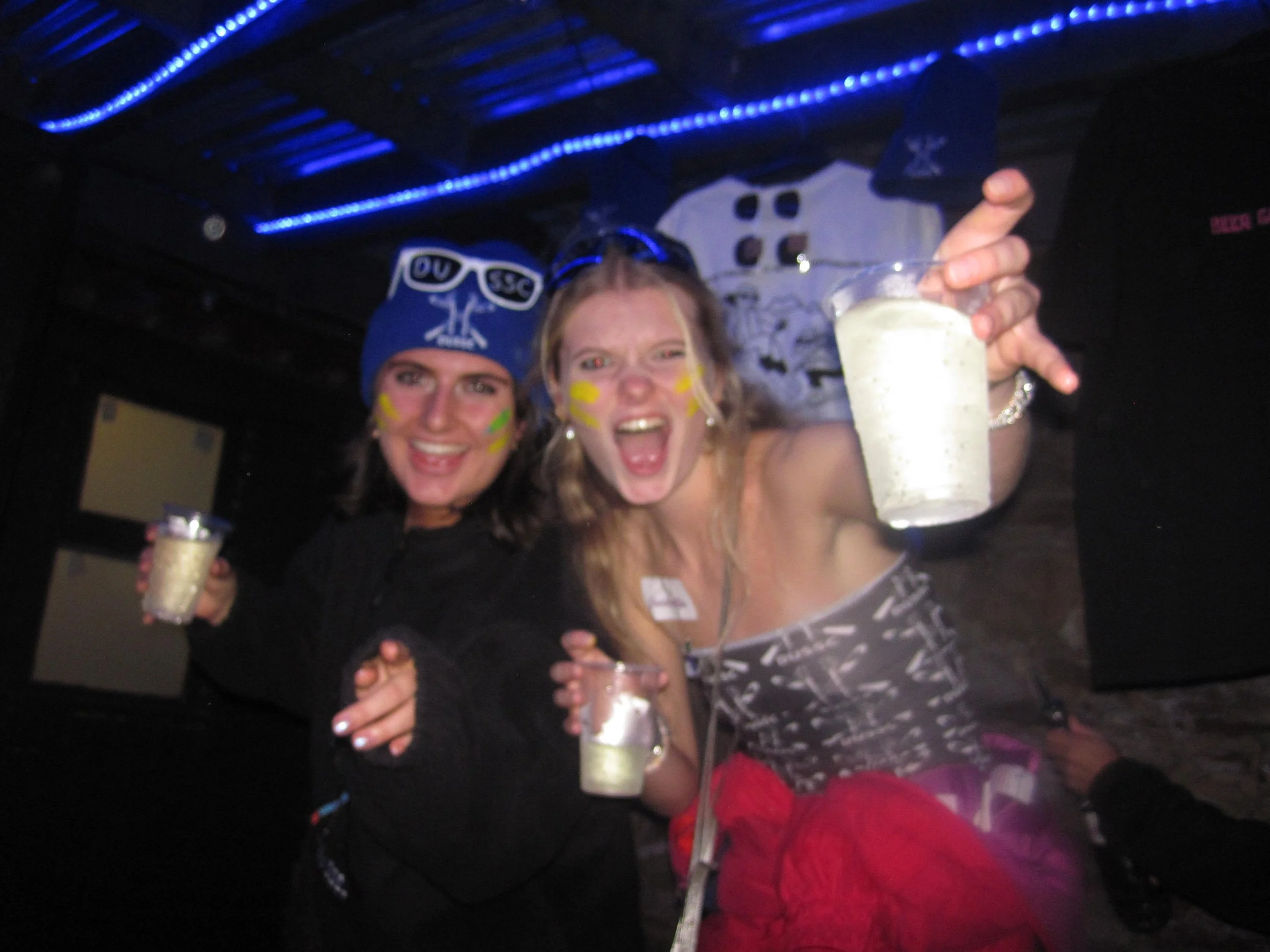 Two women celebrating, wearing face paint, holding drinks, and smiling at a party with dark lighting and blue LED lights.