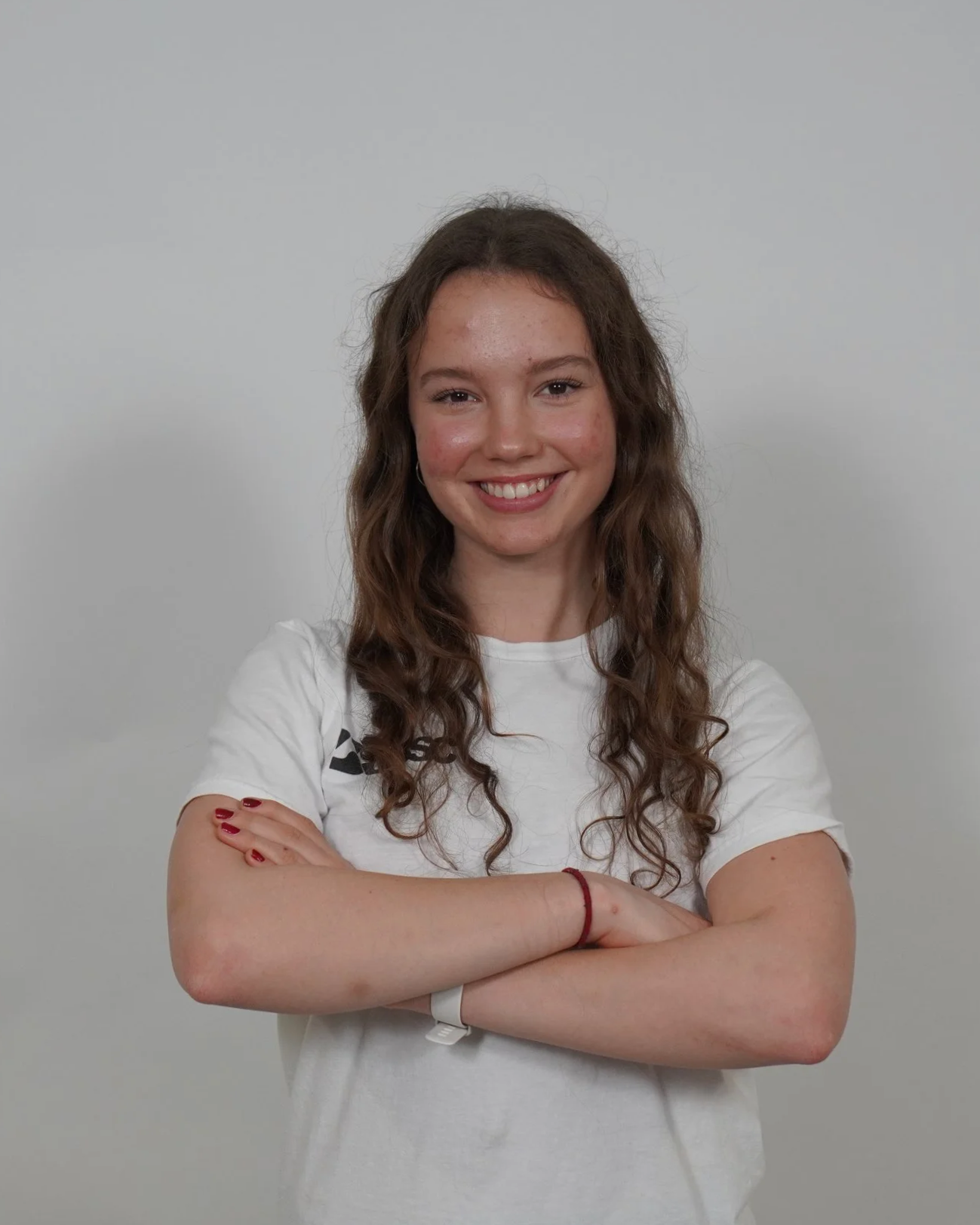 Young woman with brown curly hair smiling with arms crossed, wearing a white t-shirt and standing against a plain gray background.